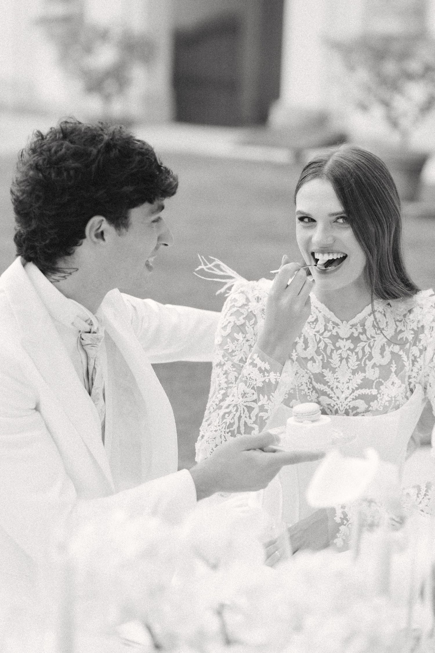 Black and white photo of a couple in white wedding attire sharing cake and laughing together at a reception. Villa Mosconi Bertani
