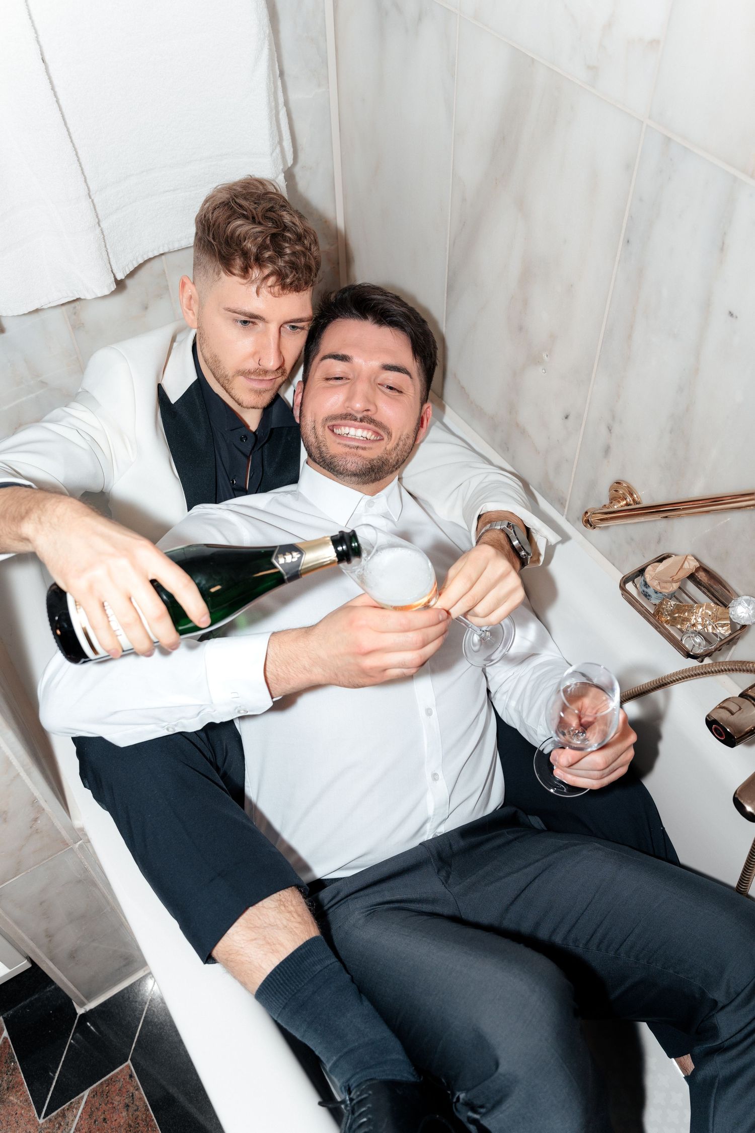 Two men in formal attire pour champagne while relaxing and laughing together in a casual setting.