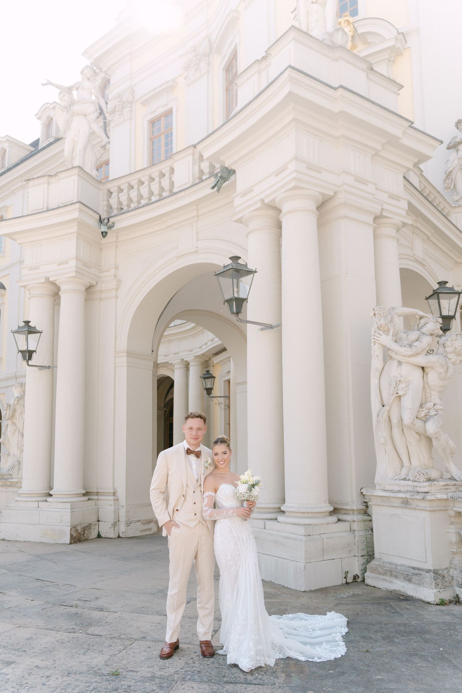 Elegant wedding couple poses in front of ornate white classical architecture with columns and statues. Schloss Ludwigsburg