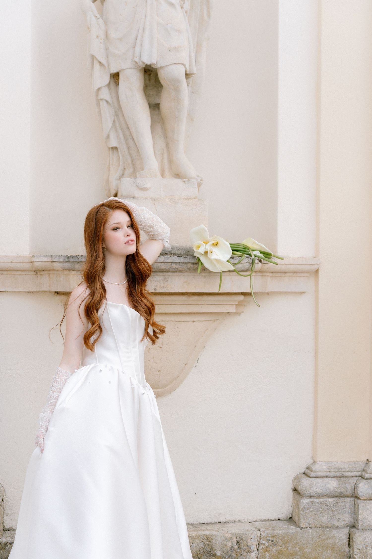Woman in flowing white dress poses gracefully against ornate stone architecture with white flowers.
