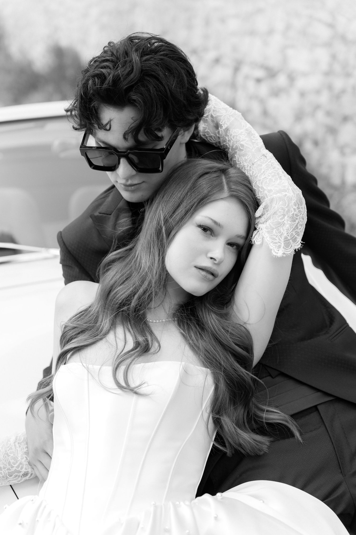 A romantic black and white portrait of a couple in formal attire posing against a car.