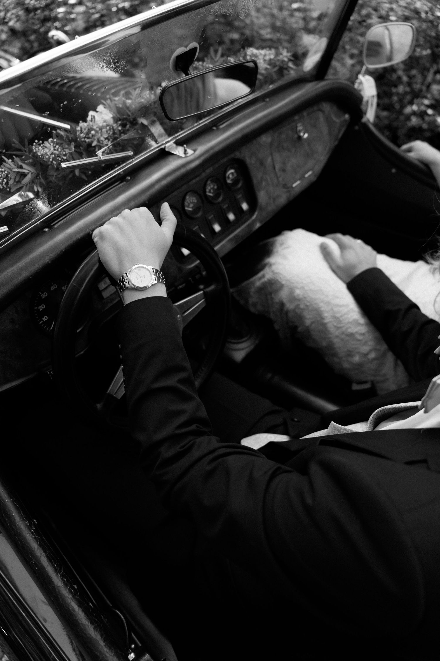 Black and white photo of a groom in a dark suit and wristwatch driving a vintage convertible, with a bride in white beside him.