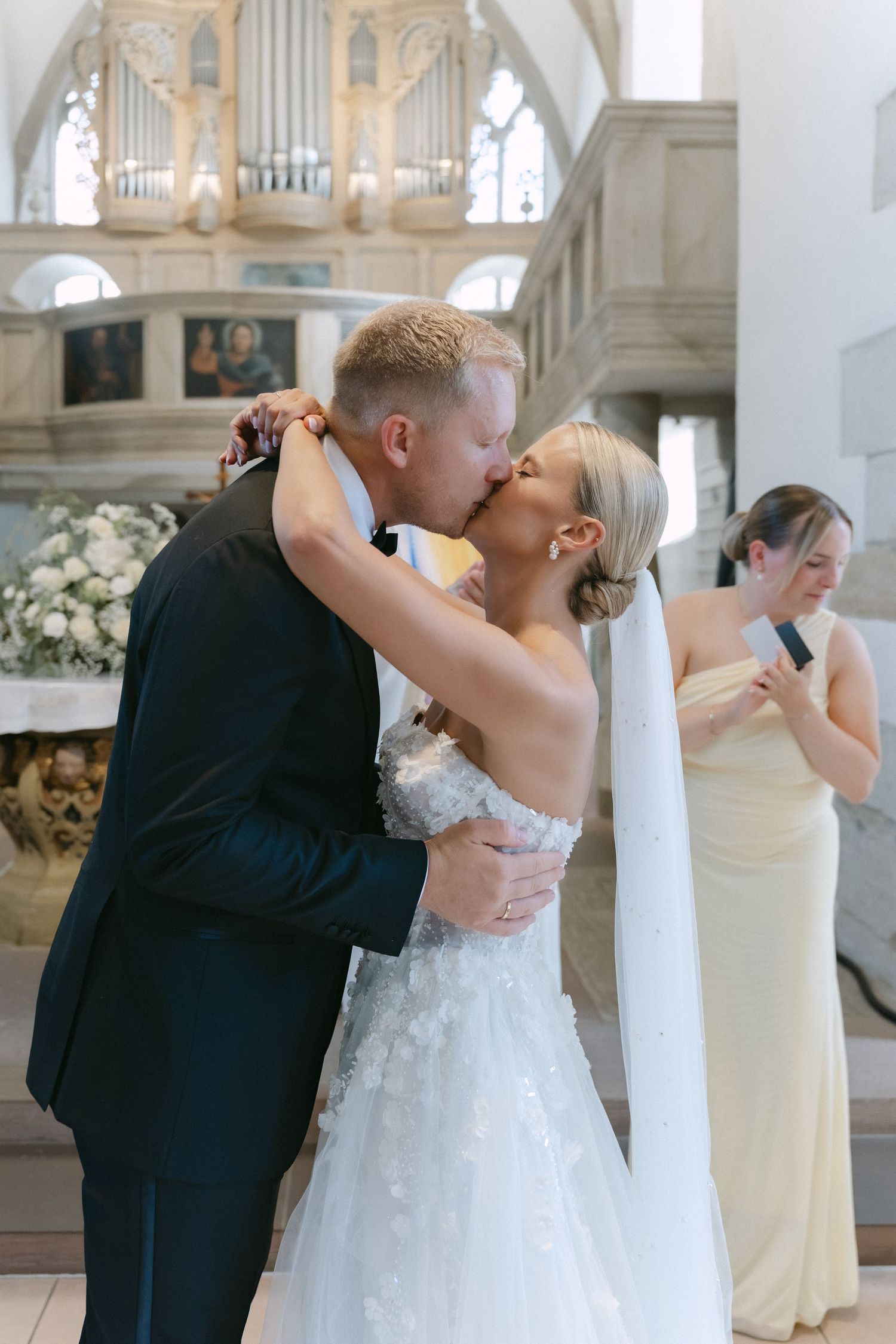 Bride in a lace wedding gown and groom in a dark suit share a kiss at a church altar as a bridesmaid in yellow looks on.