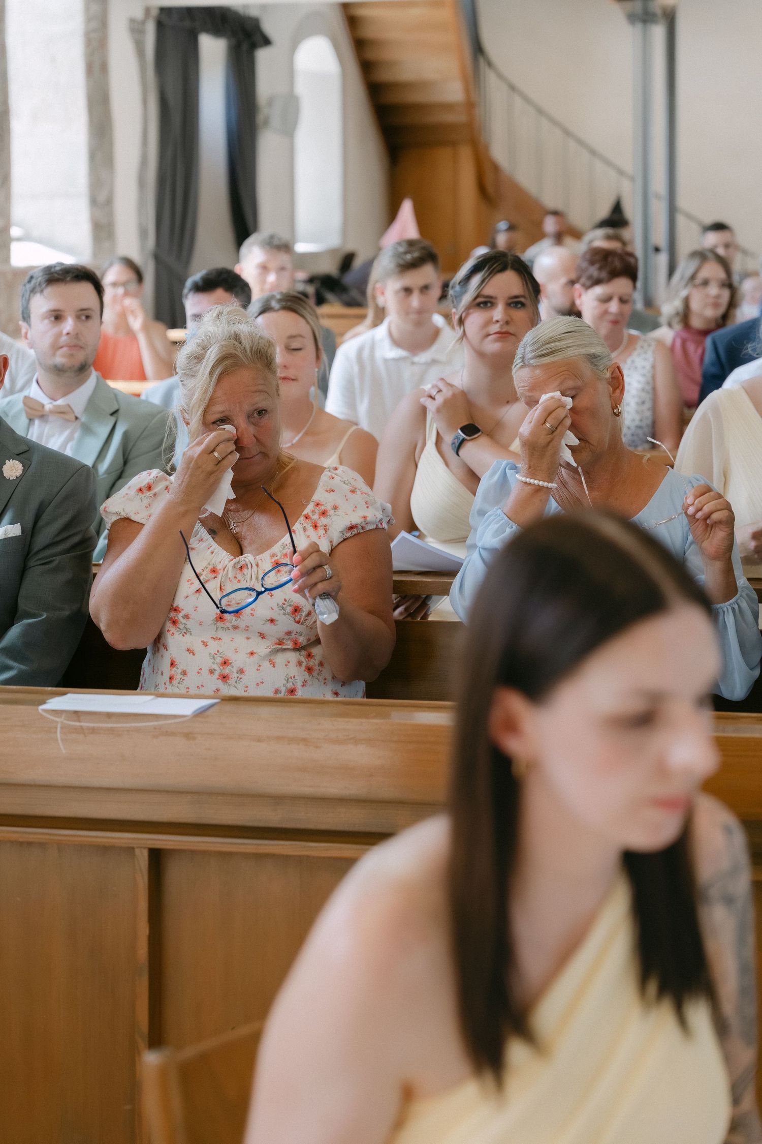 Emotional wedding guests dab tears with tissues while seated in wooden church pews during a ceremony.