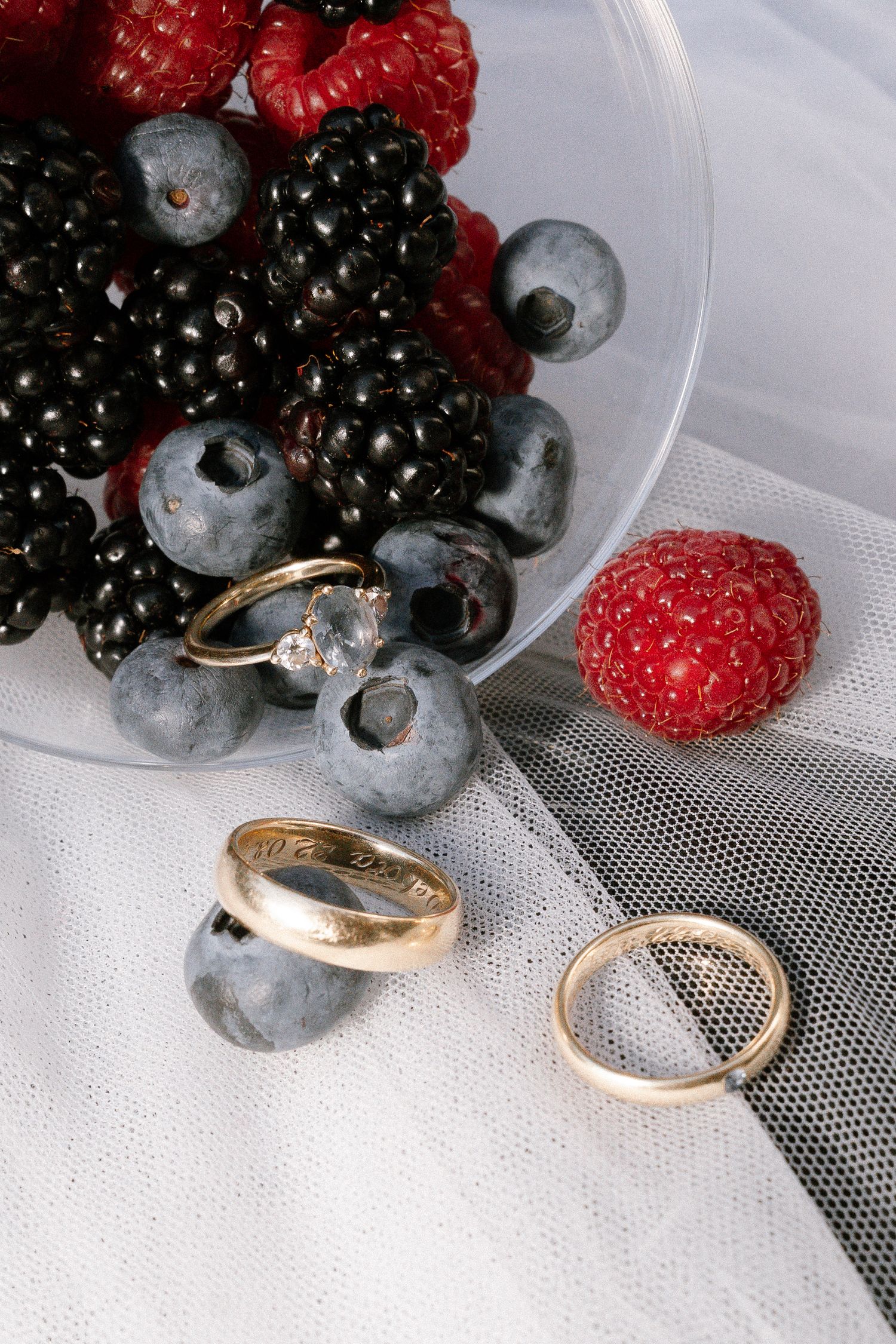 Gold wedding rings and a diamond engagement ring rest on white tulle beside a glass bowl of blueberries, blackberries and raspberries.