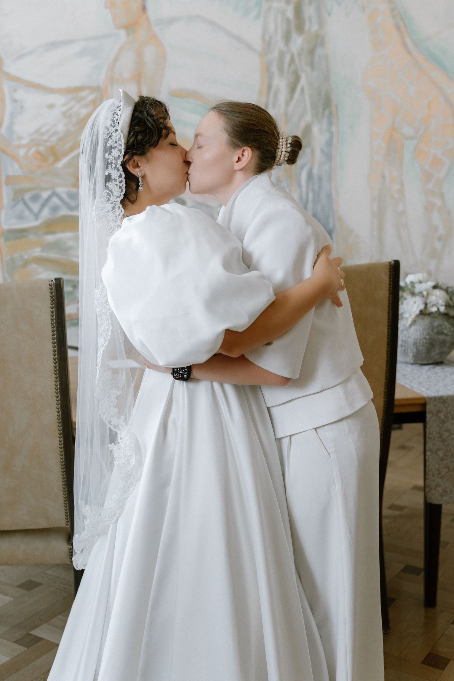 Two brides in white wedding attire share a romantic kiss, one wearing a lace veil, in an elegantly decorated room.