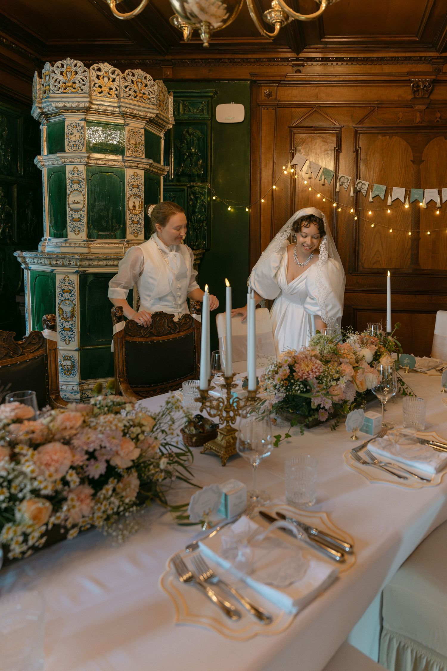 Bride and wedding planner arranging candles and floral centerpieces on elegant reception table in wood-paneled venue.