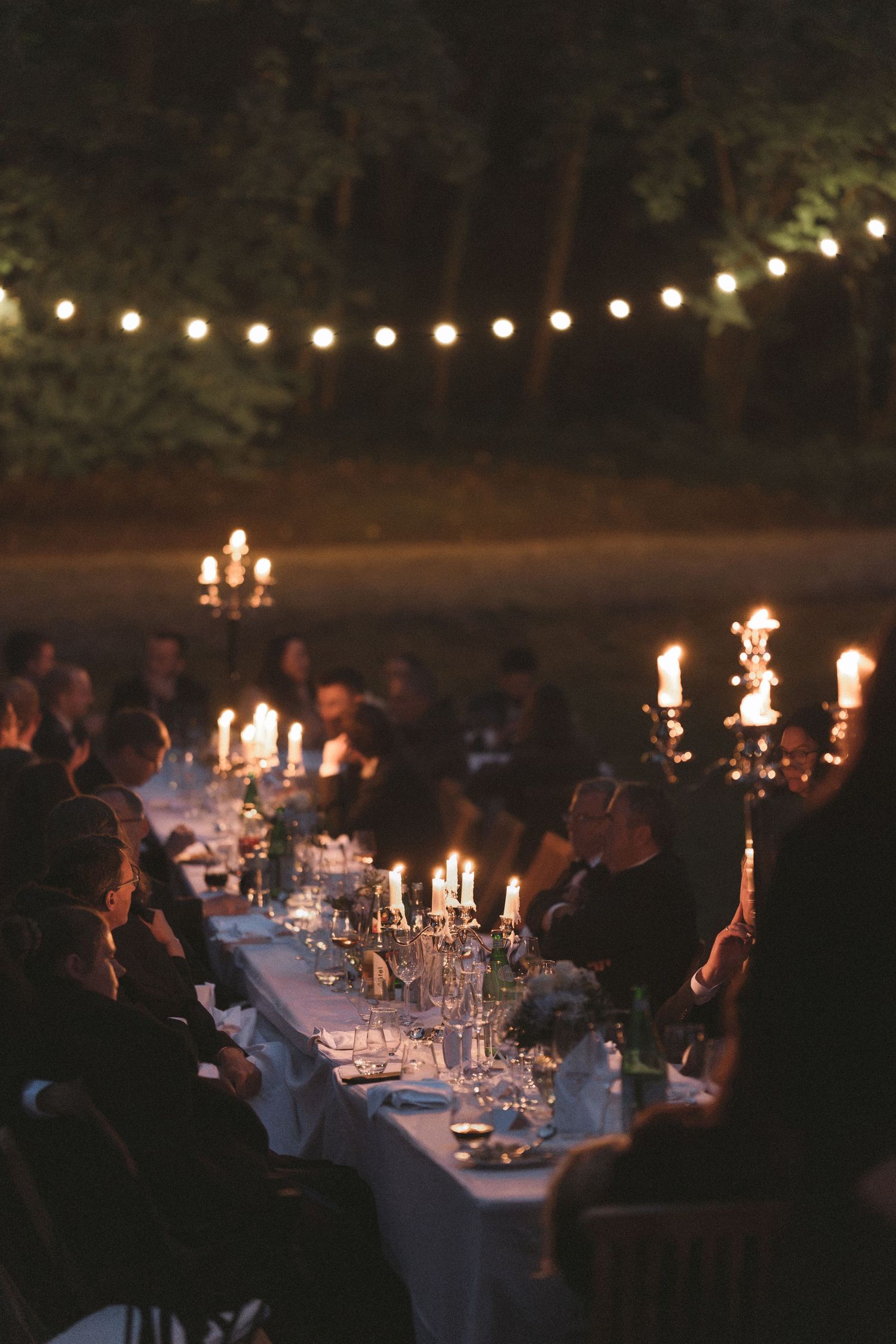 Guests dine at a long candlelit table with candelabras and glassware in a wooded outdoor setting under glowing string lights at dusk.