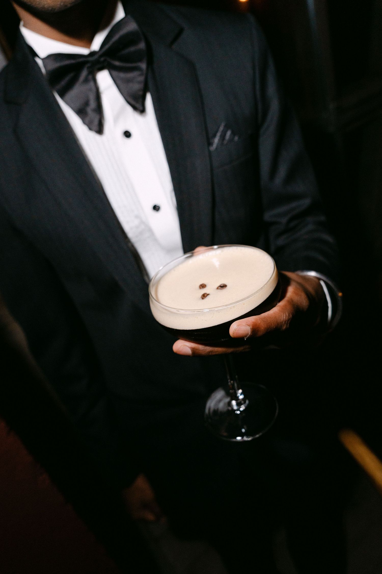 Bartender in a black tuxedo and bow tie holds an espresso martini topped with creamy foam and three coffee beans in a coupe glass.