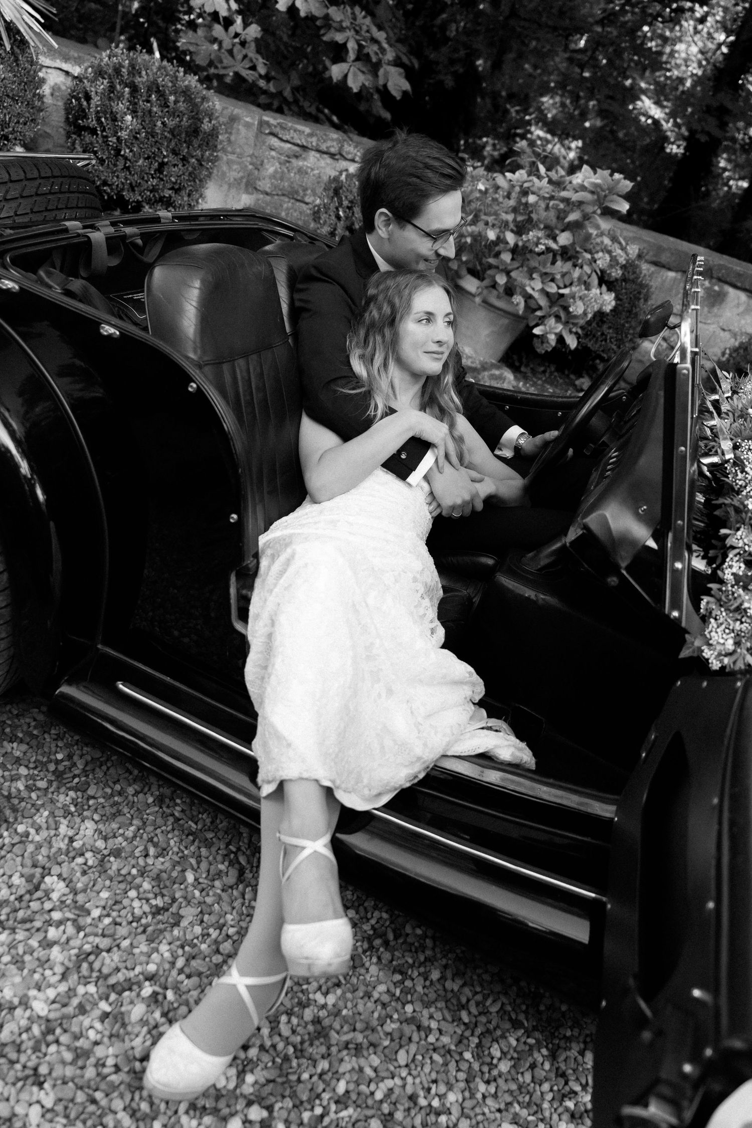 Black and white photo of a bride in a lace dress and a groom in glasses relaxing together in a vintage convertible car.
