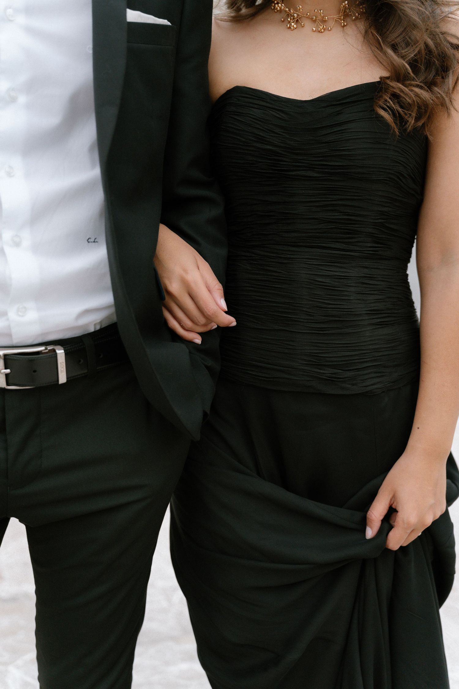 Couple in elegant black formal wear, woman in strapless ruched gown, man in dark suit, standing close together.