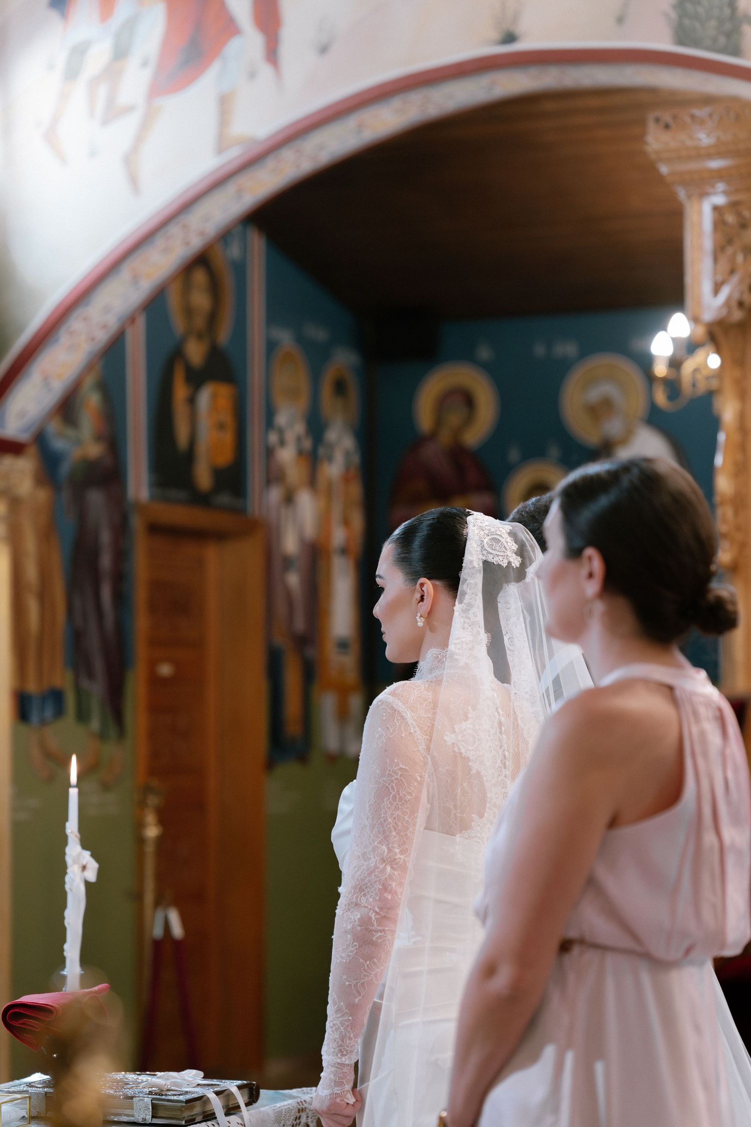 Bride in lace wedding dress and veil stands before Orthodox church icons during ceremony.