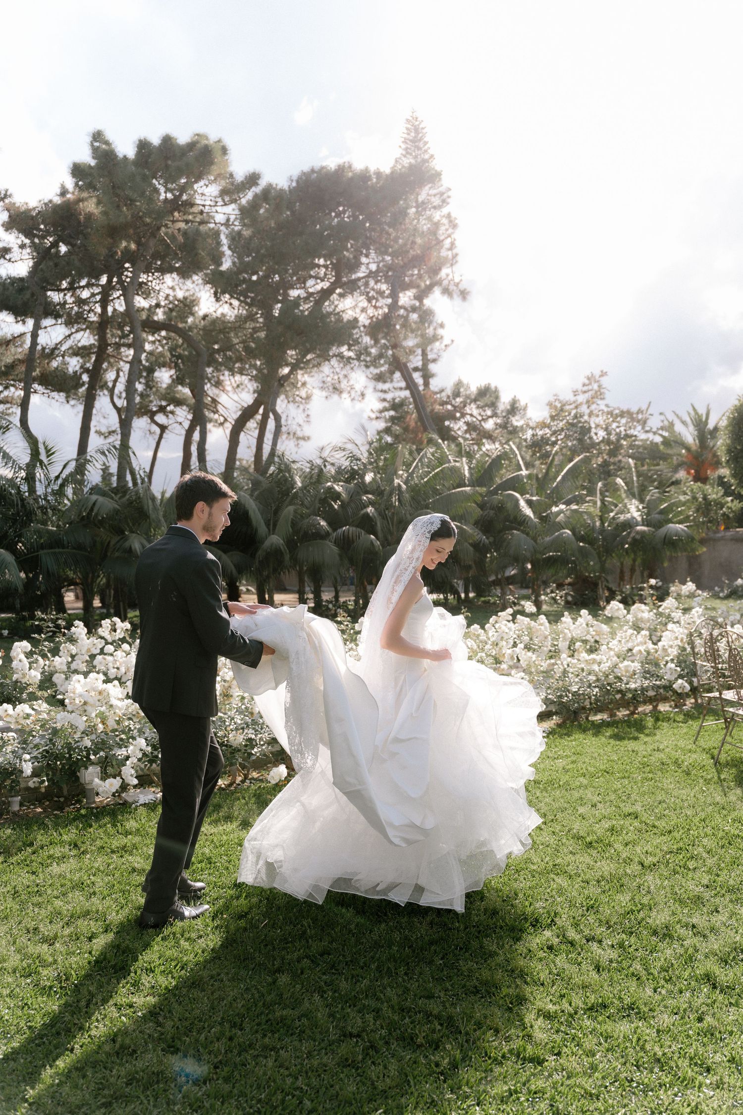 Bride in flowing white gown and veil dances with groom in black suit on green lawn surrounded by white flowers.