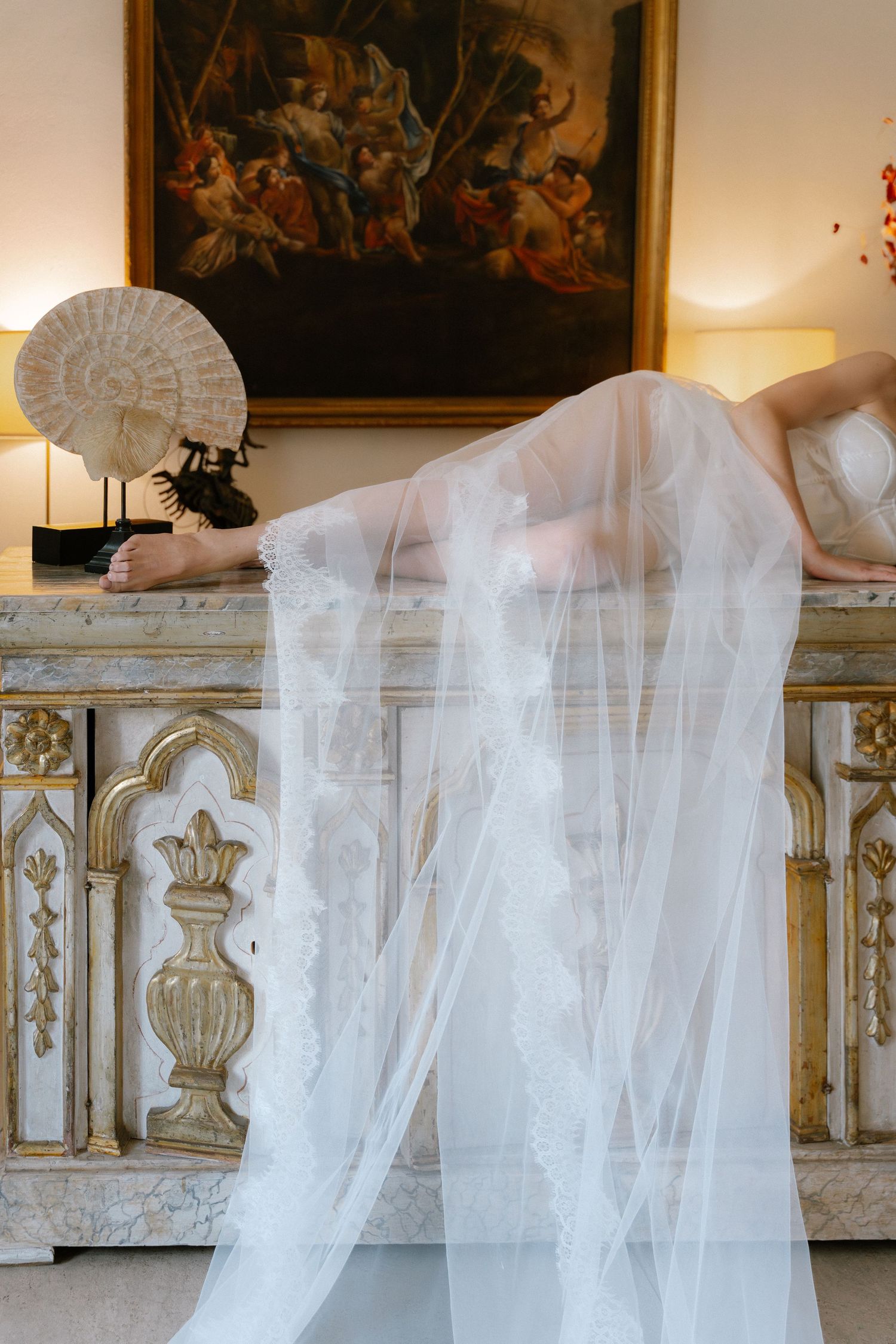 Bride in white lace wedding dress and veil draped over ornate vintage dresser with classical painting backdrop.