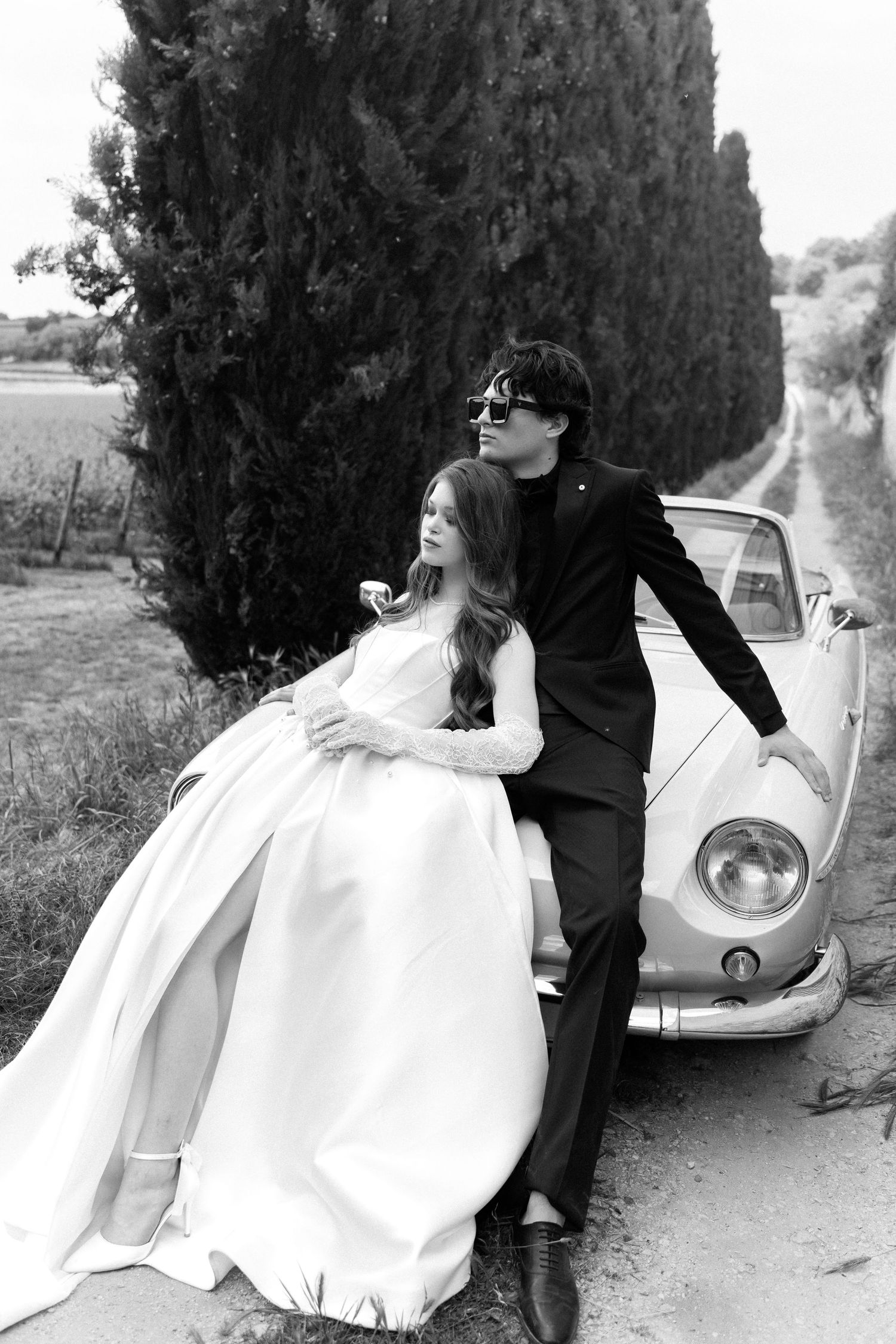 A romantic black and white wedding photo series showing a couple posing beside a vintage car in a countryside setting.