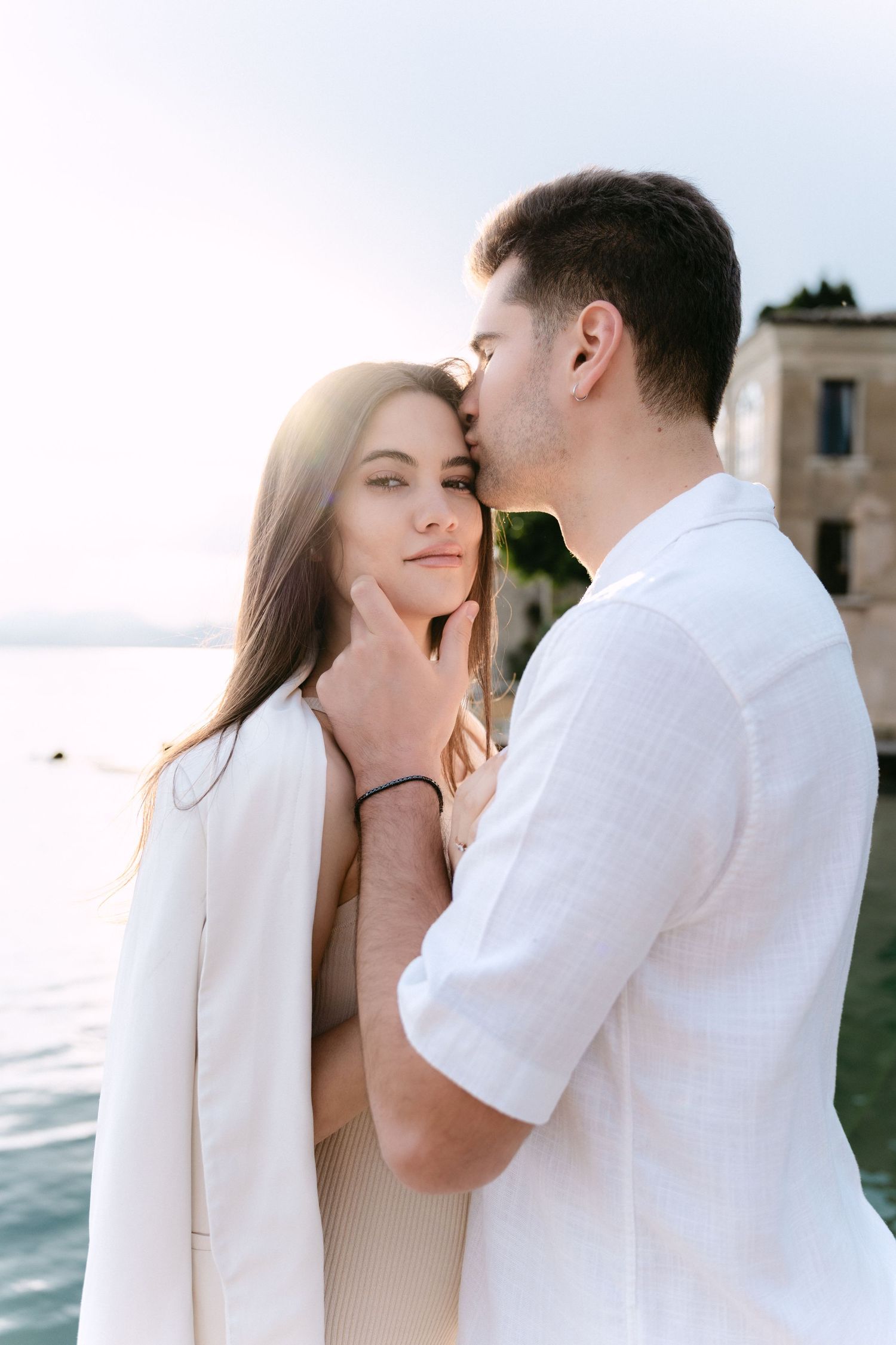 Romantic couple in white outfits sharing an intimate moment by the water during sunset.