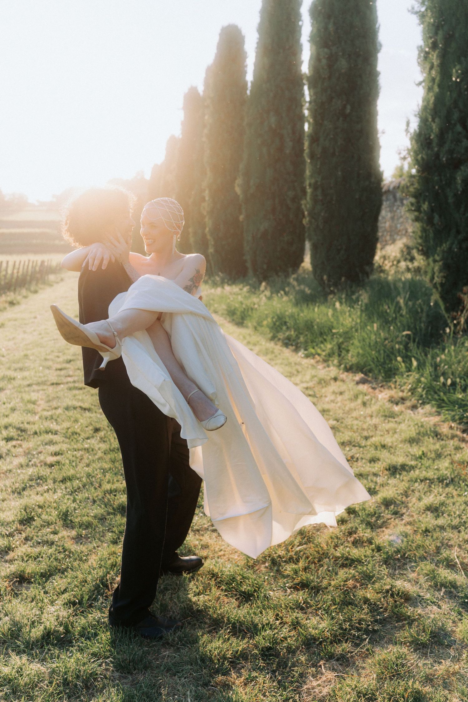 A romantic couple in wedding attire dances together in a sun-drenched cypress tree-lined path during golden hour.