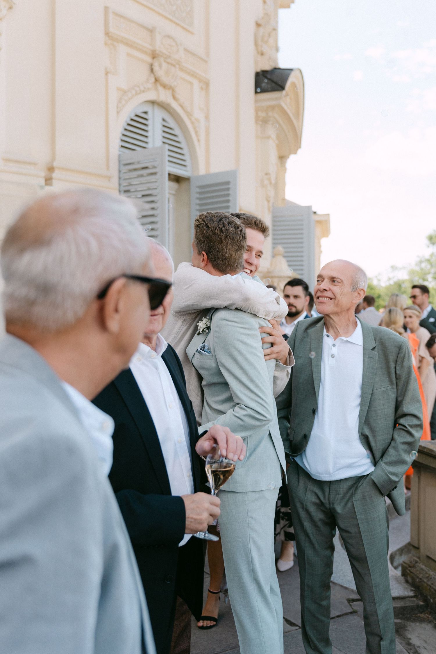 A group of well-dressed guests in suits gather at an outdoor event, sharing embraces and conversation. Schloss Solitude