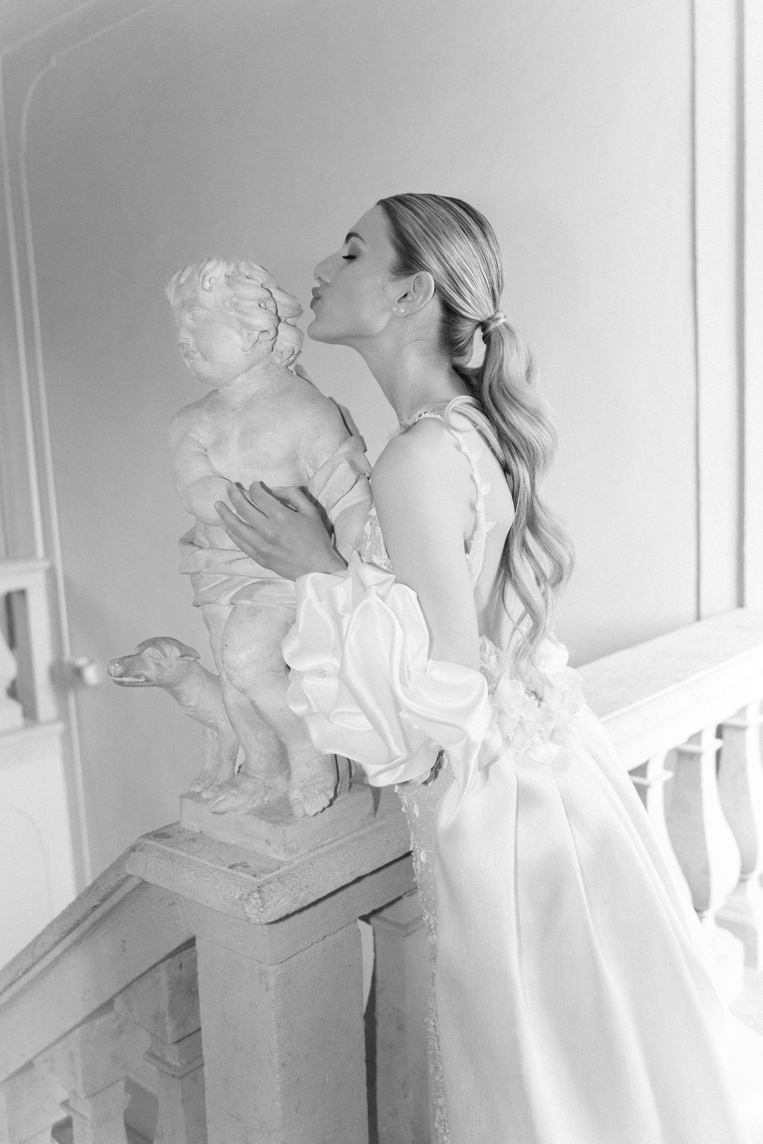 A woman in an elegant white dress kisses a cherub statue in a romantic black and white photograph. Wedding Verona