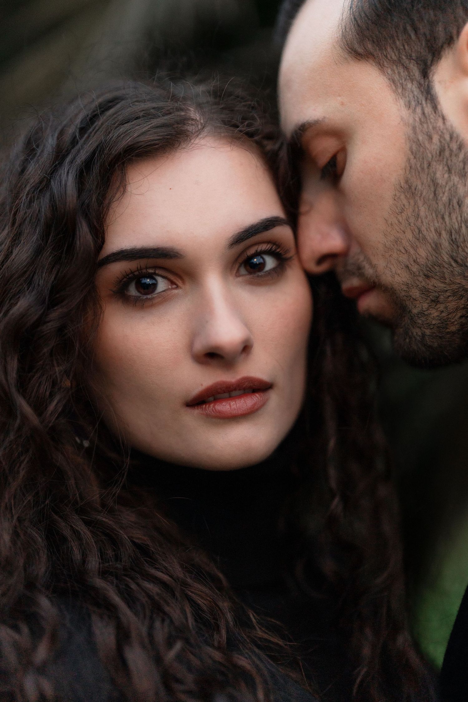 Intimate romantic portrait of a couple in a dark moody setting with curly dark hair and soft lighting.