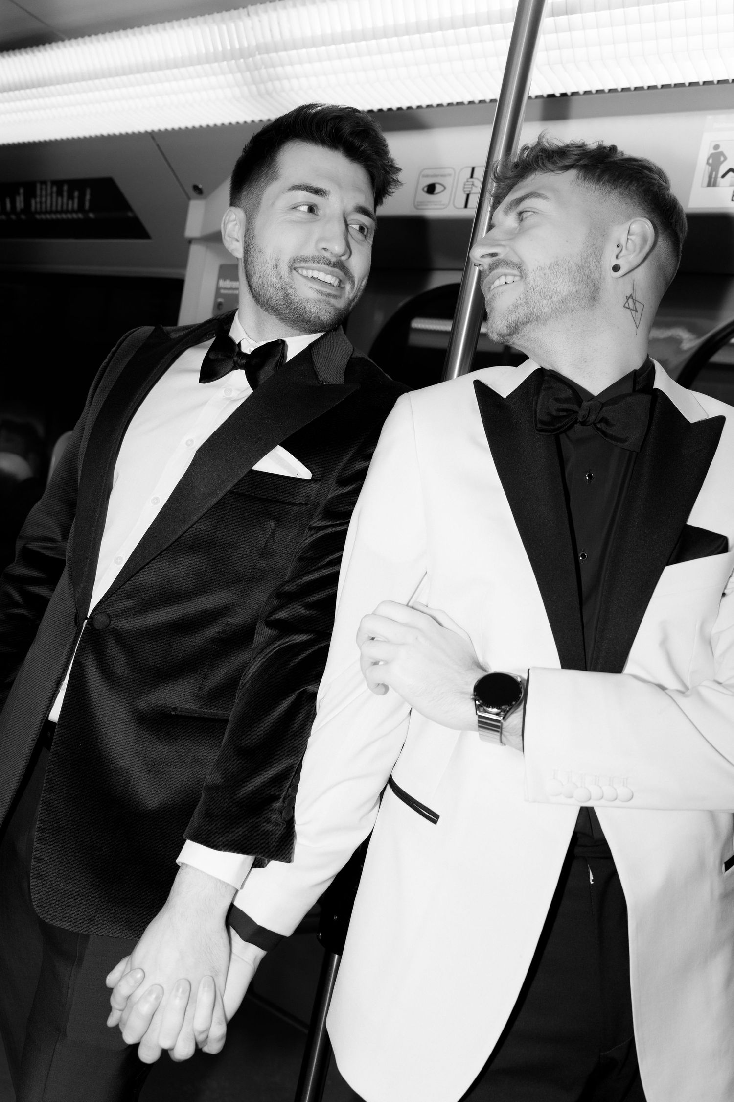 Two men at formal event wearing contrasting black and white tuxedos share a moment together in black and white photo.