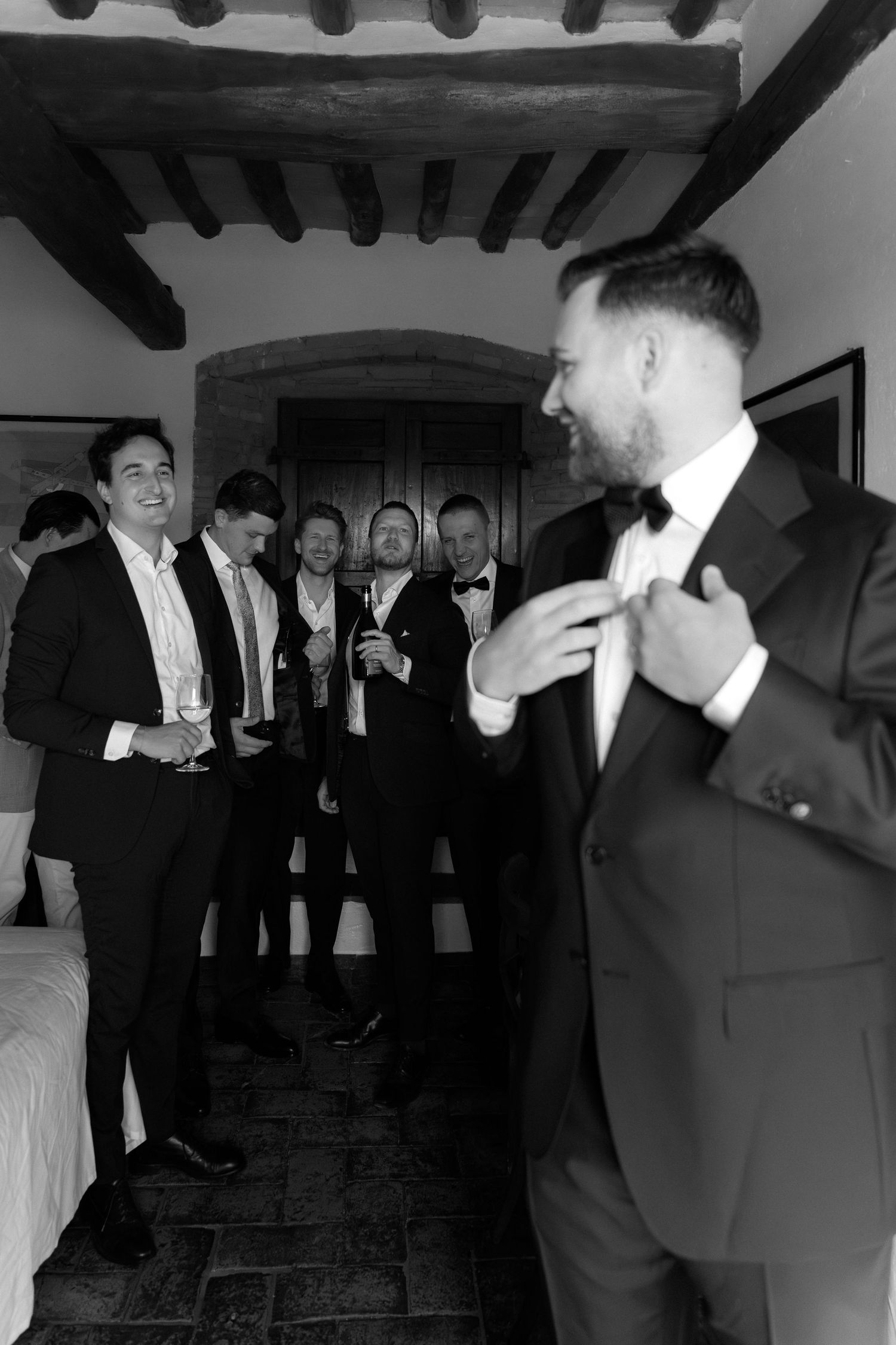 Black and white photo of groom adjusting bow tie while groomsmen smile and celebrate in rustic venue.