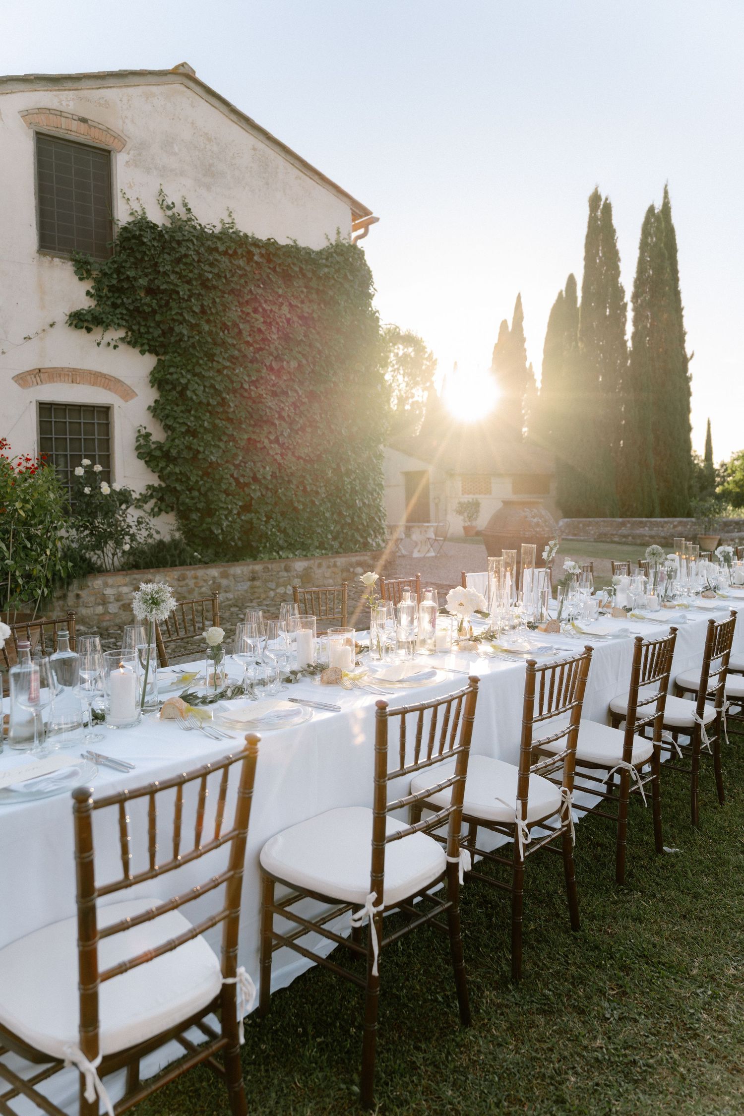 Elegant outdoor wedding reception with white-draped long tables and chiavari chairs at golden hour near an ivy-covered villa.