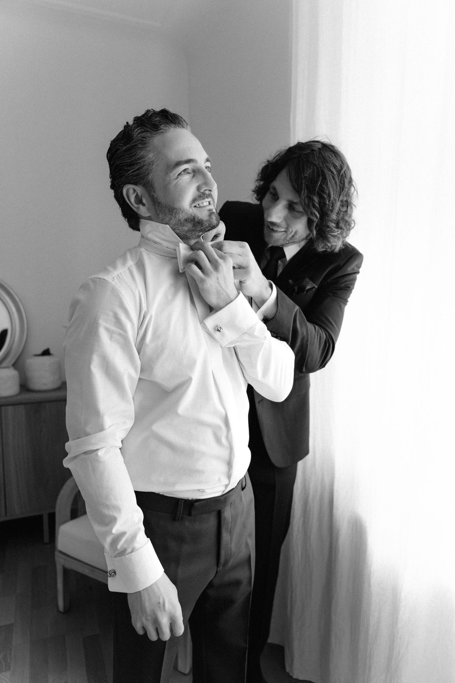 Black and white photo of a woman helping a smiling groom adjust his bow tie before the wedding ceremony.