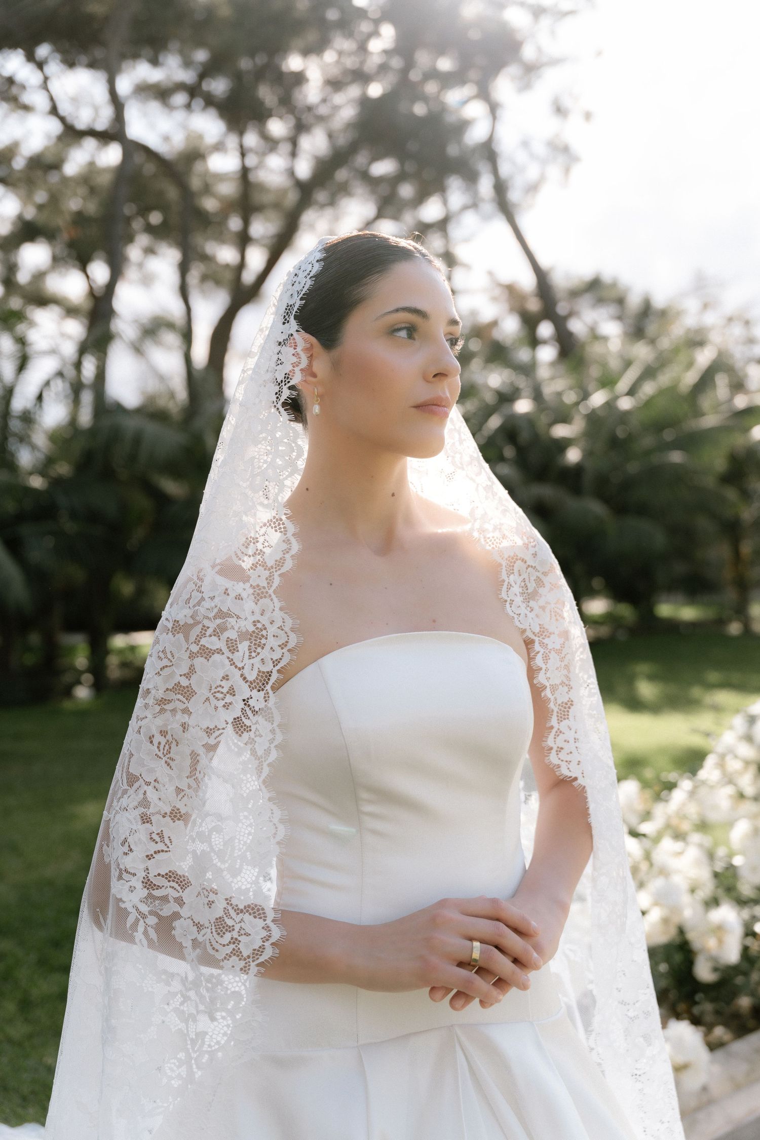 Bride in strapless white gown and lace veil poses outdoors with lush greenery, exuding elegance on her wedding day.