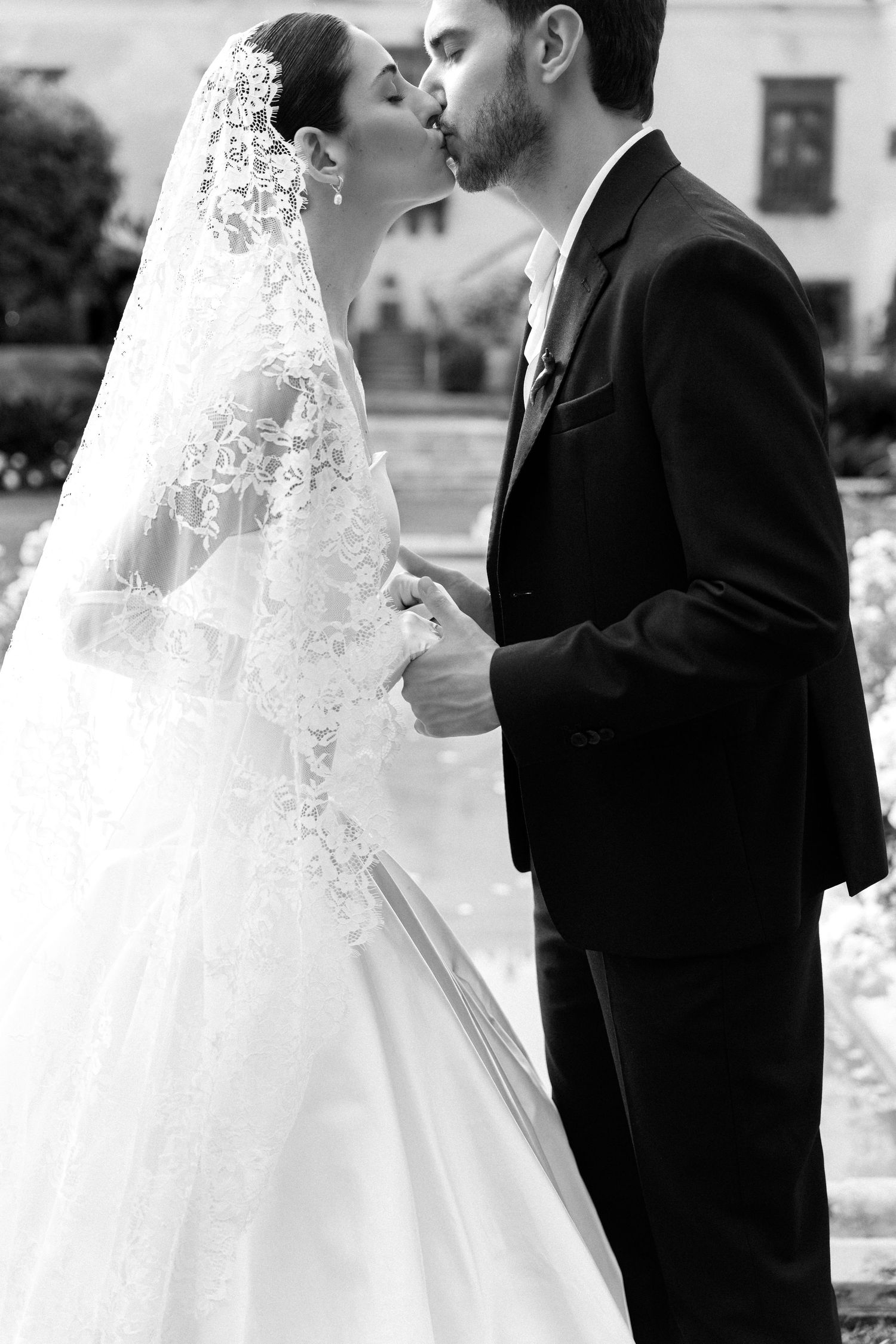 Black and white photo of a bride and groom sharing a kiss, bride wearing lace veil and gown, groom in dark suit.
