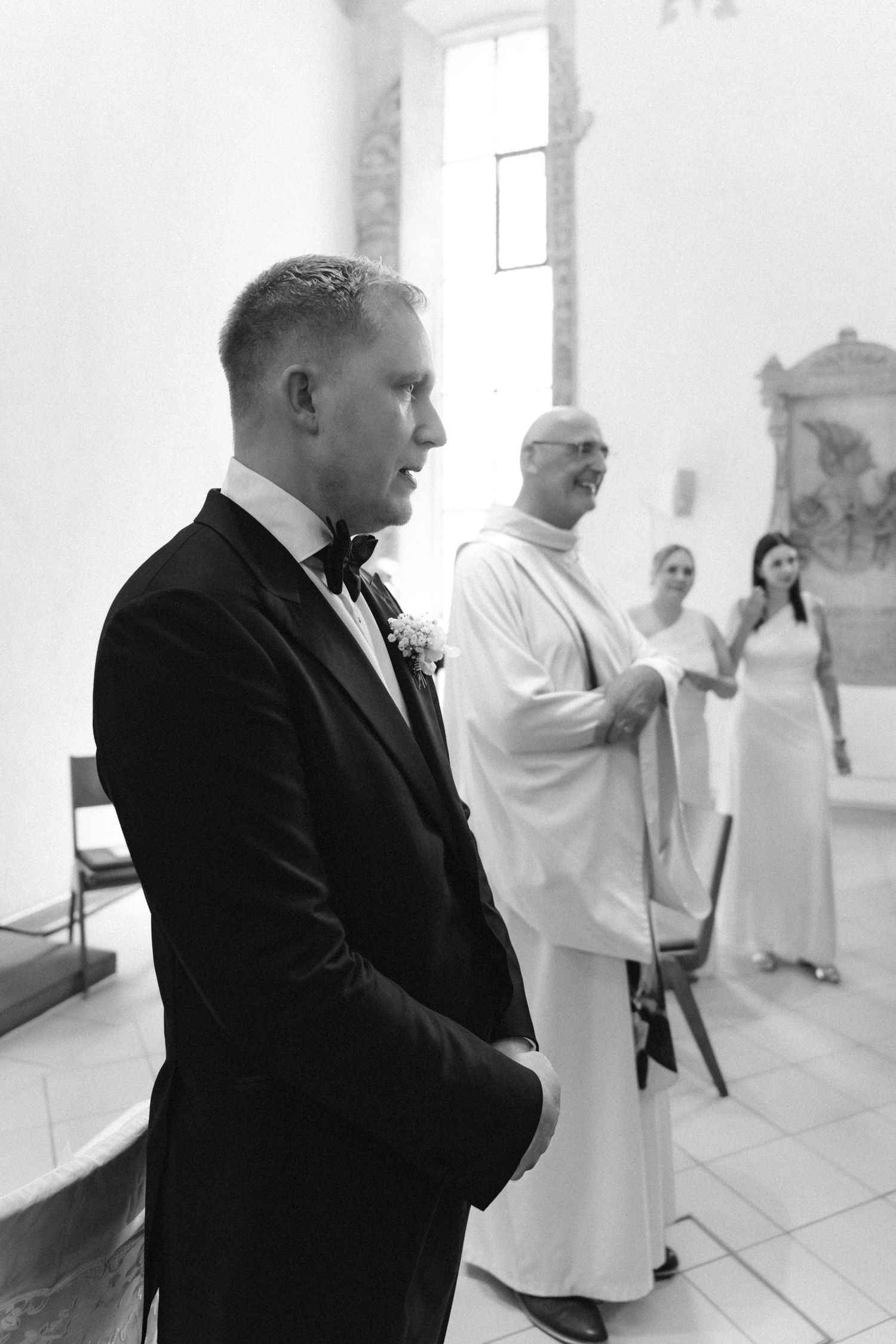 Black and white photo of a groom in a tuxedo and bow tie waiting at the altar as a priest and wedding guests look on in a church.