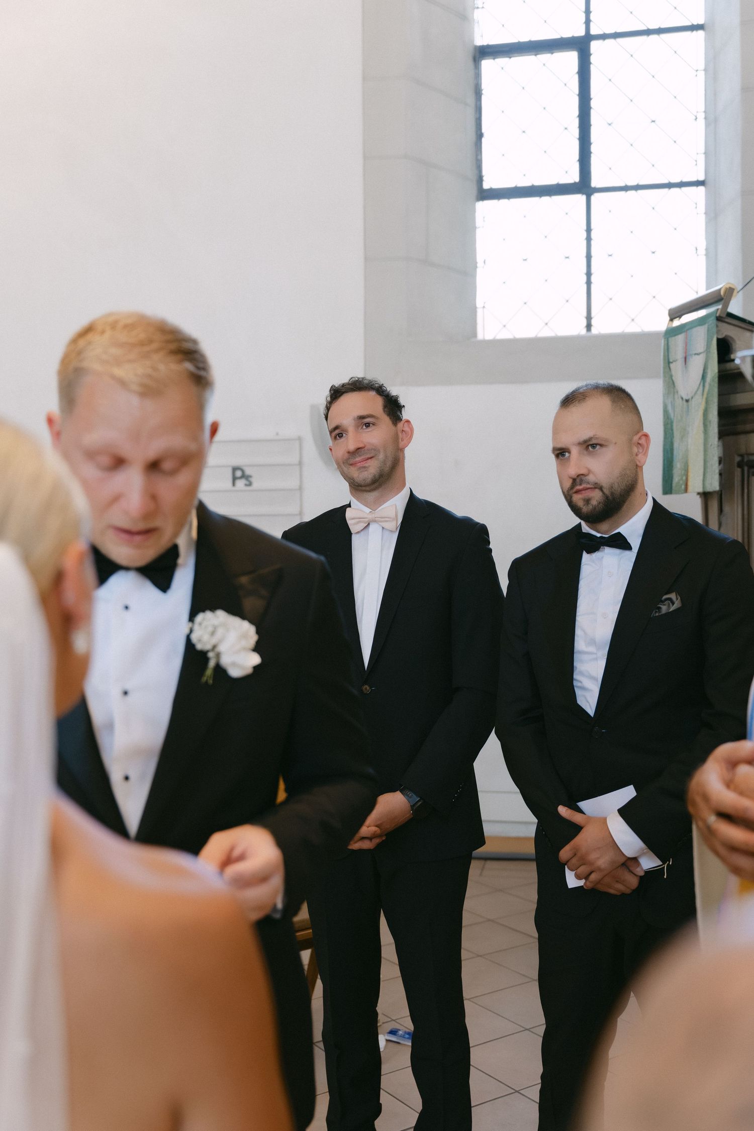 Groomsmen in black tuxedos and bow ties stand smiling beside the groom during a wedding ceremony in a bright stone church.