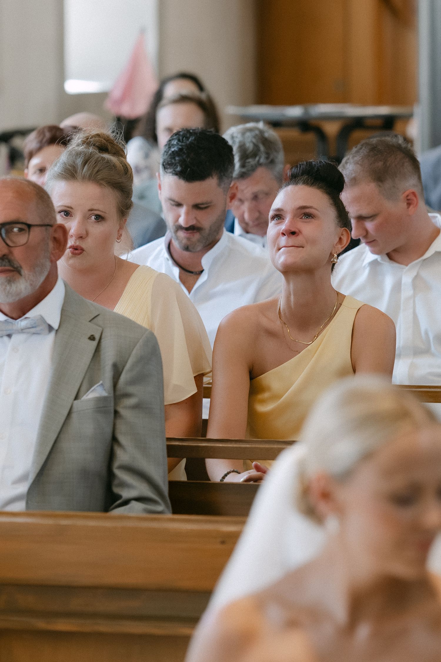 Wedding guests in formal attire sit on wooden church pews, watching the ceremony with attentive and emotional expressions.