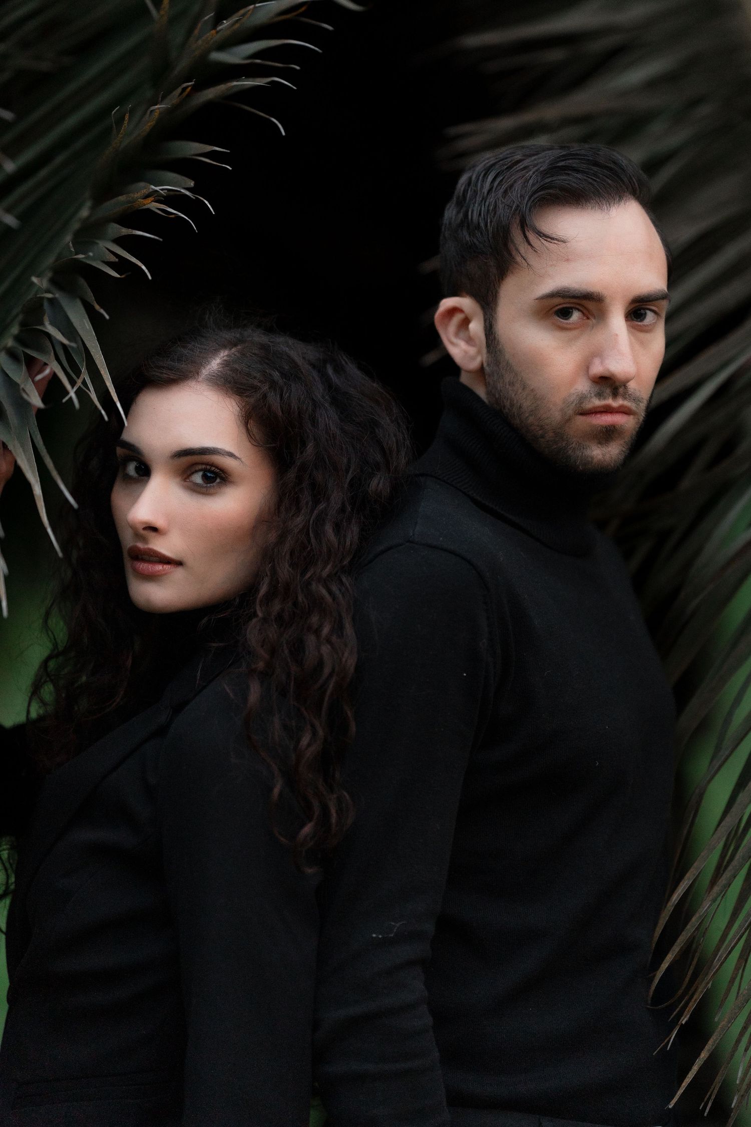 Two people in black turtleneck sweaters pose artistically against tropical foliage in a moody portrait.