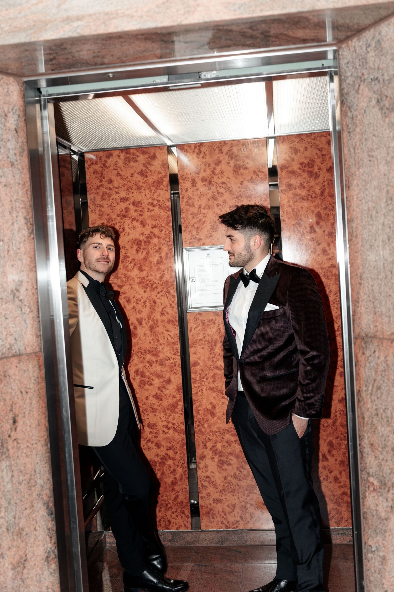 Two men in formal tuxedos standing in an elevator with ornate copper-colored walls.