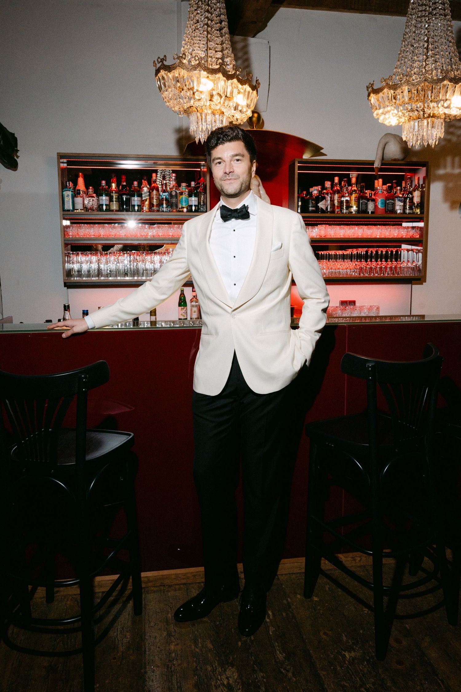 A person in a white tuxedo jacket and black bow tie poses at a bar with crystal chandeliers overhead.