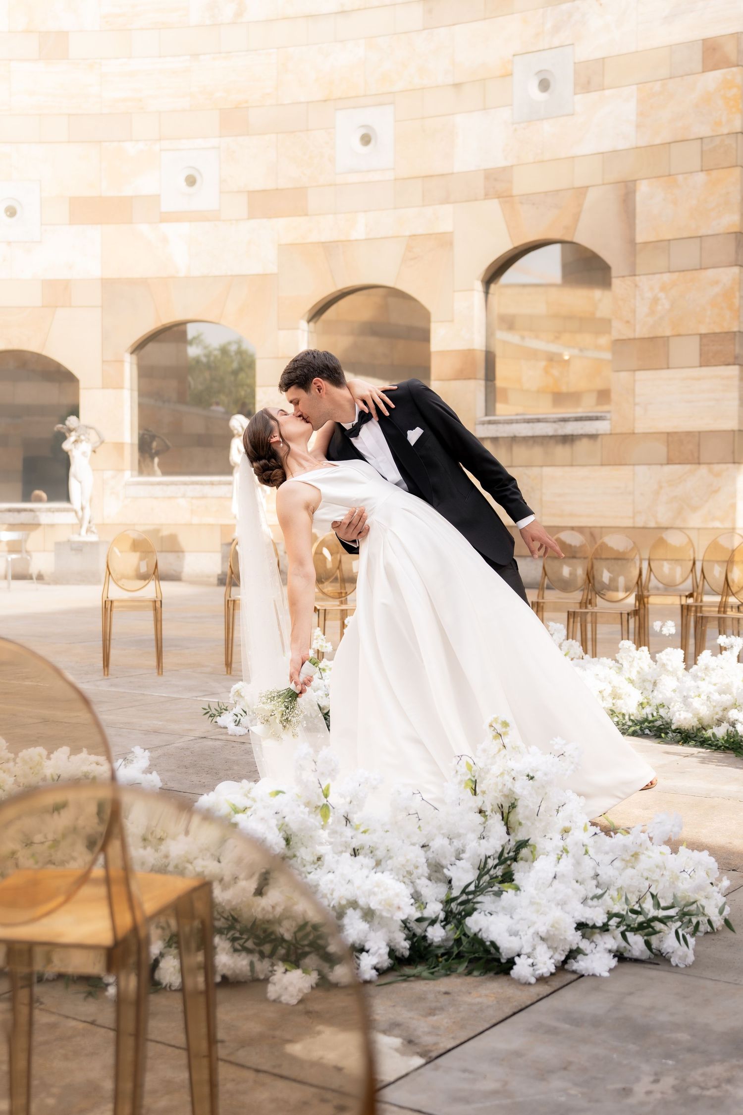 A romantic wedding scene with a couple dipping and kissing amid white floral decorations and gold chairs at a stone venue.