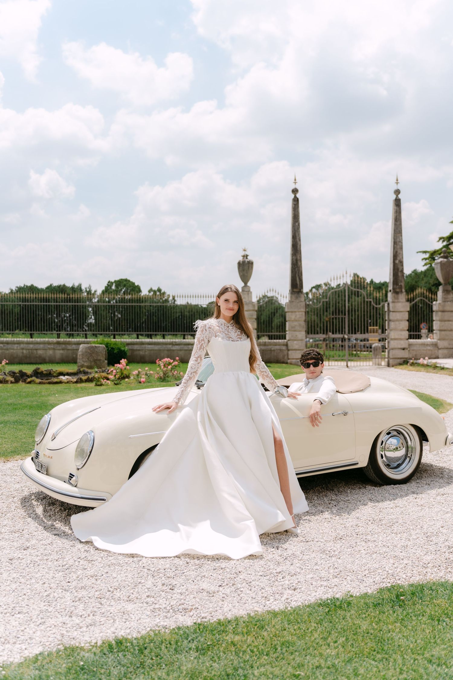 A flowing white wedding gown cascades elegantly against a vintage cream-colored luxury automobile on a gravel driveway. Villa MosconiBertani