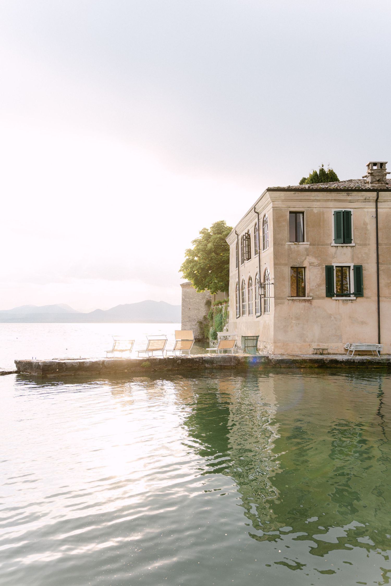 Abandoned stone buildings partially submerged in calm waters with mountains visible in the background.