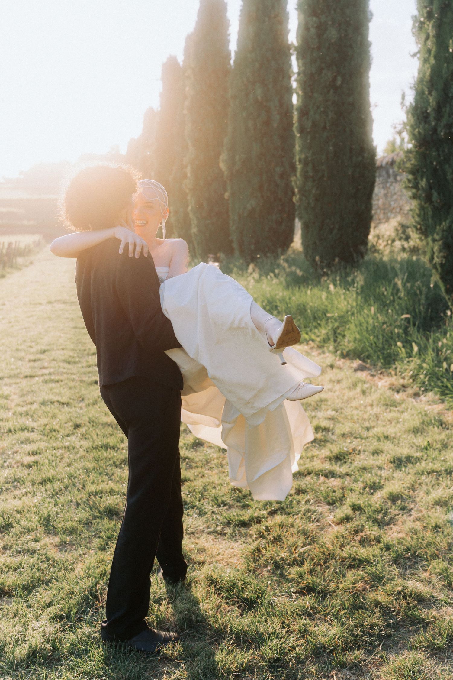 Silhouetted couple dancing in a sunlit field lined with tall cypress trees at golden hour.