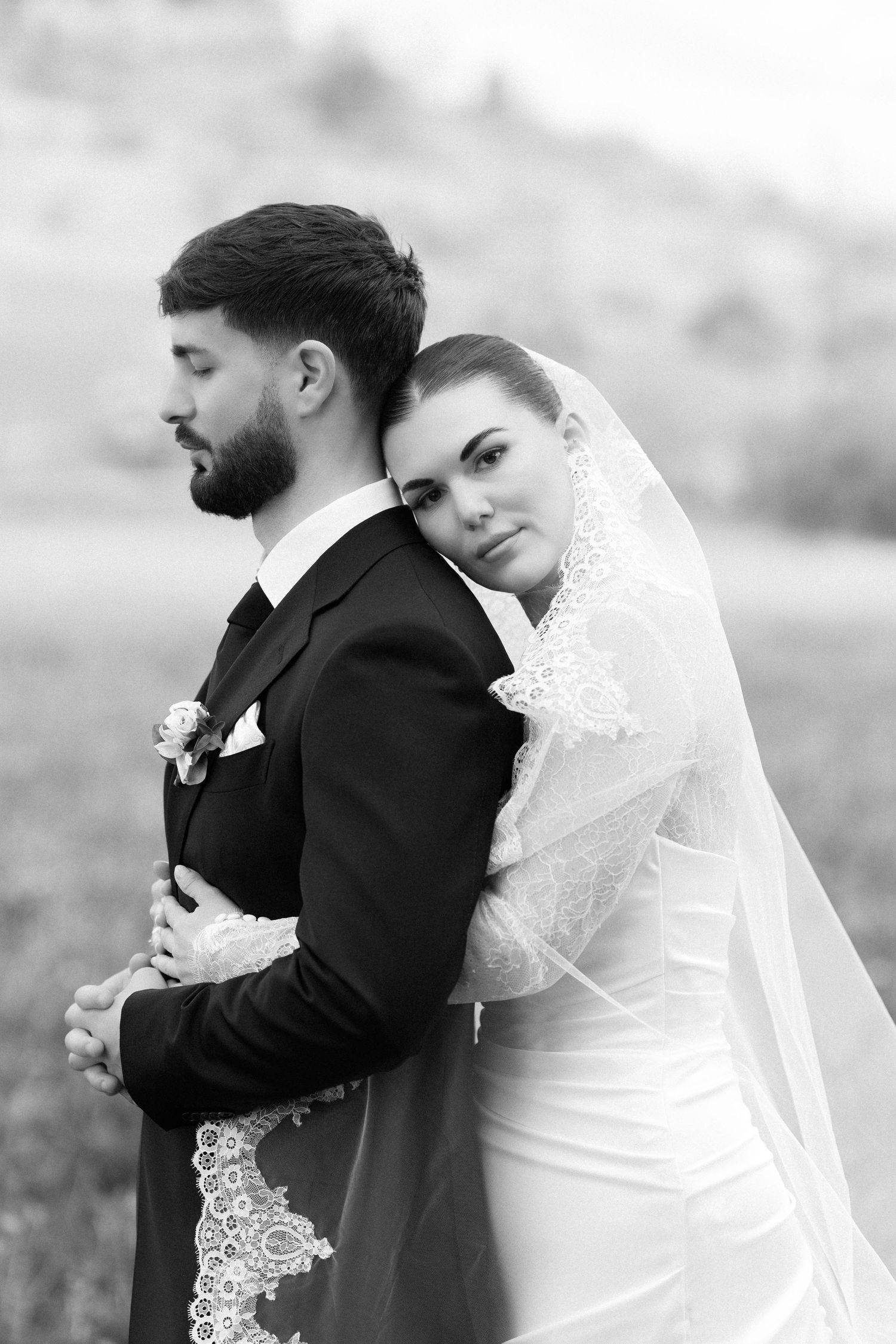 Black and white wedding photo of bride leaning on groom's back, wearing lace veil, outdoors.