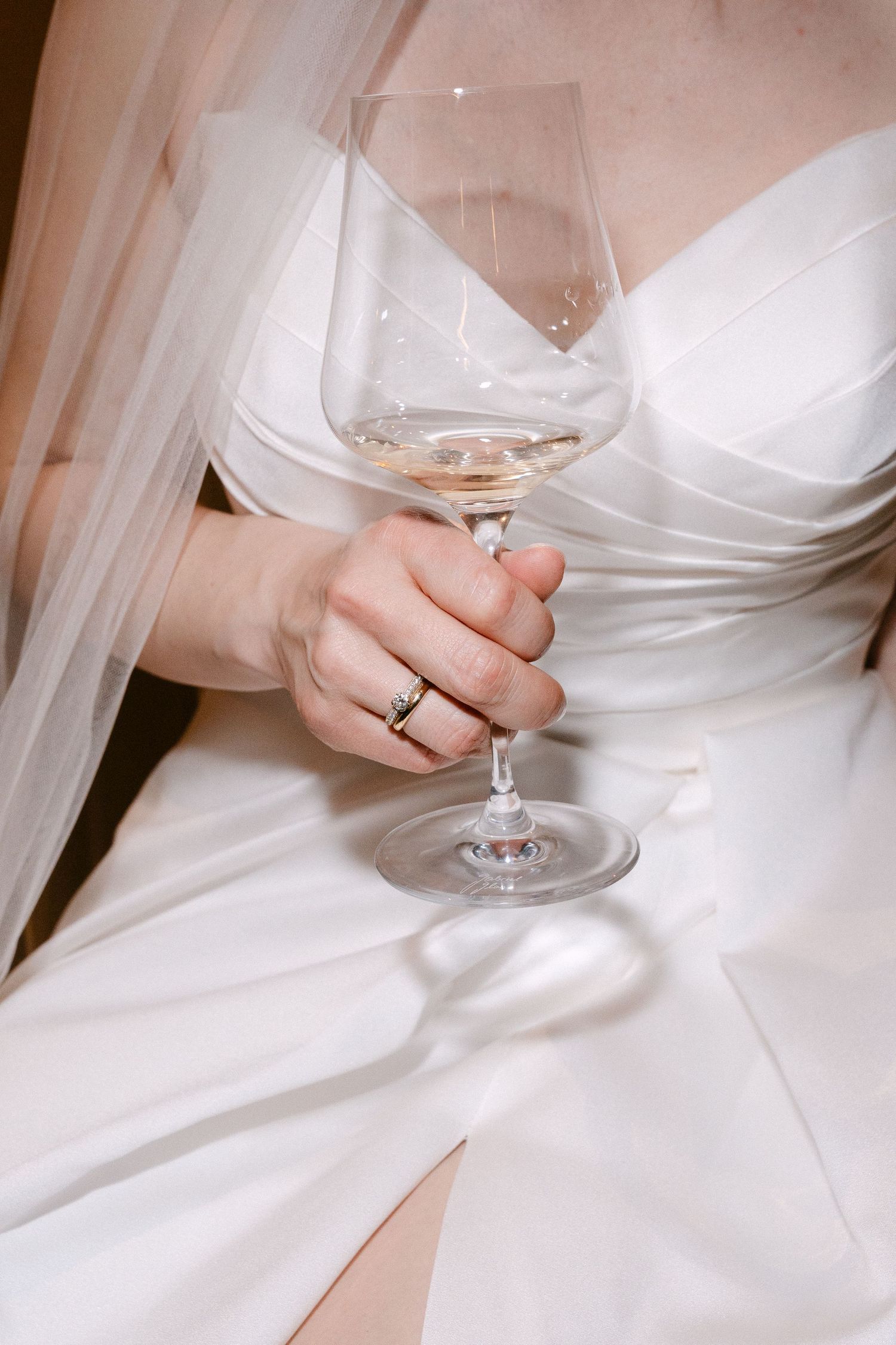 Bride in white strapless wedding gown holding a glass of white wine, with engagement ring visible on her finger.