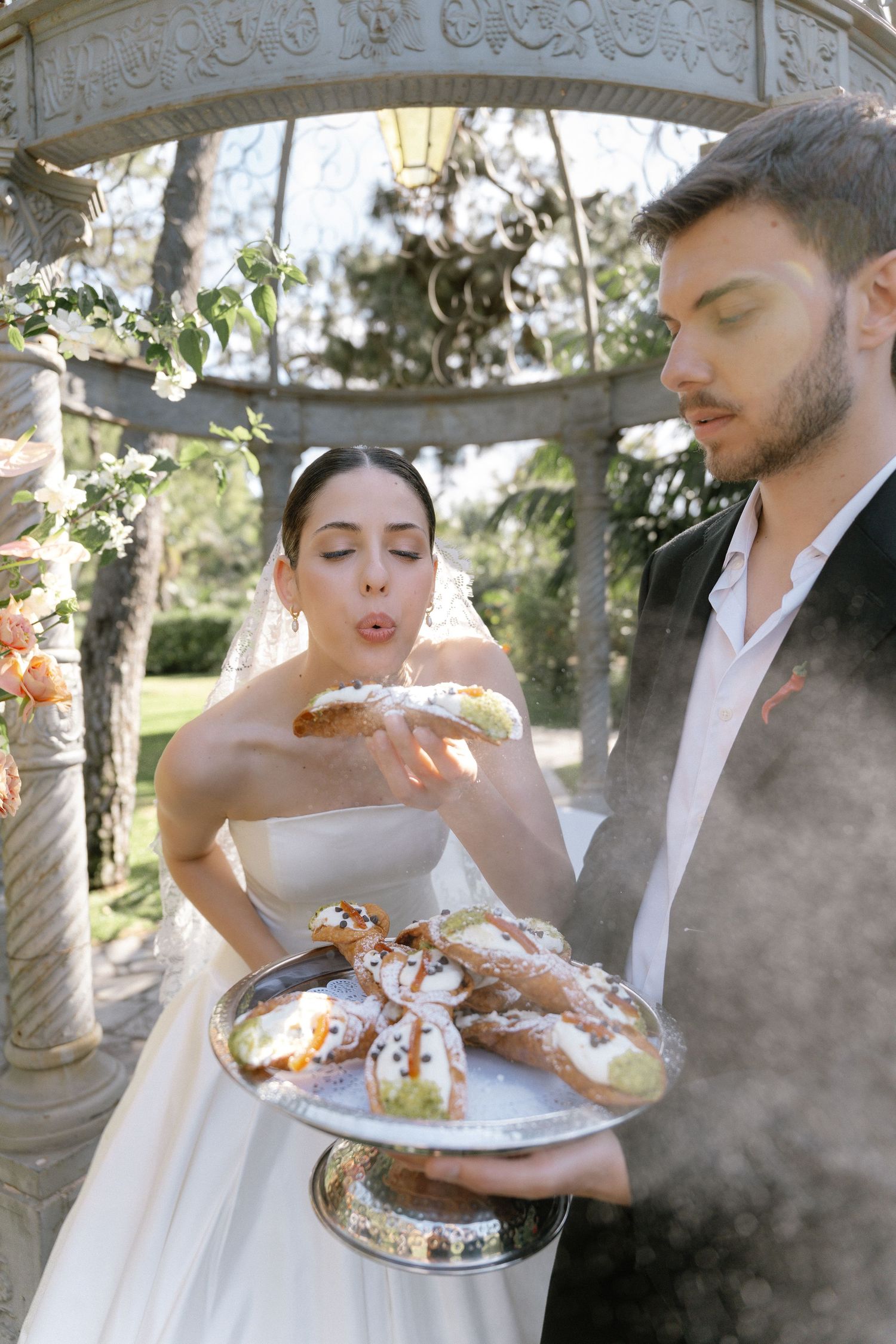 Bride blows on a pastry while groom holds a silver tray of eclairs under a stone gazebo.