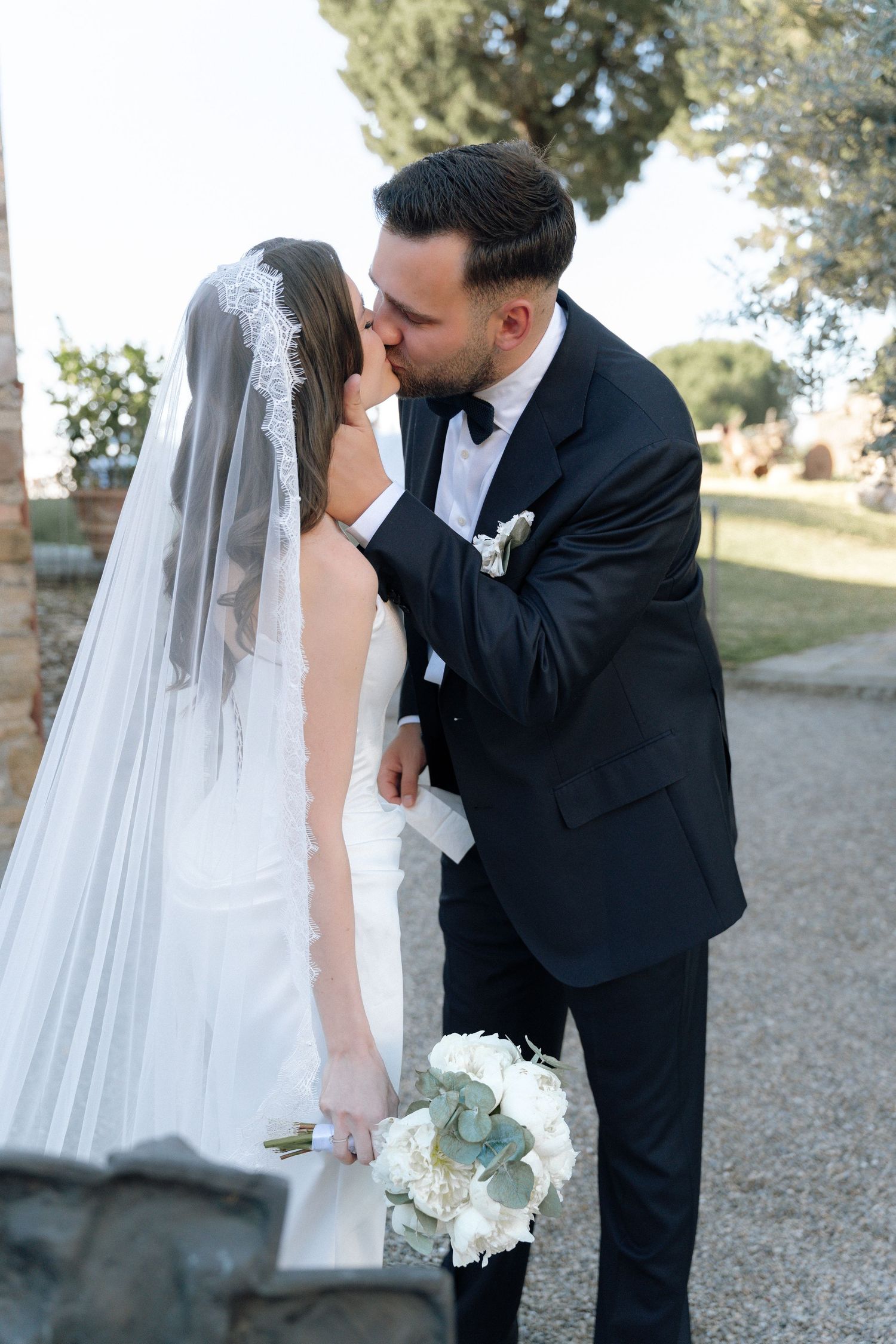 A bride and groom share a romantic kiss outdoors, she in a white gown with veil, he in a black tuxedo.