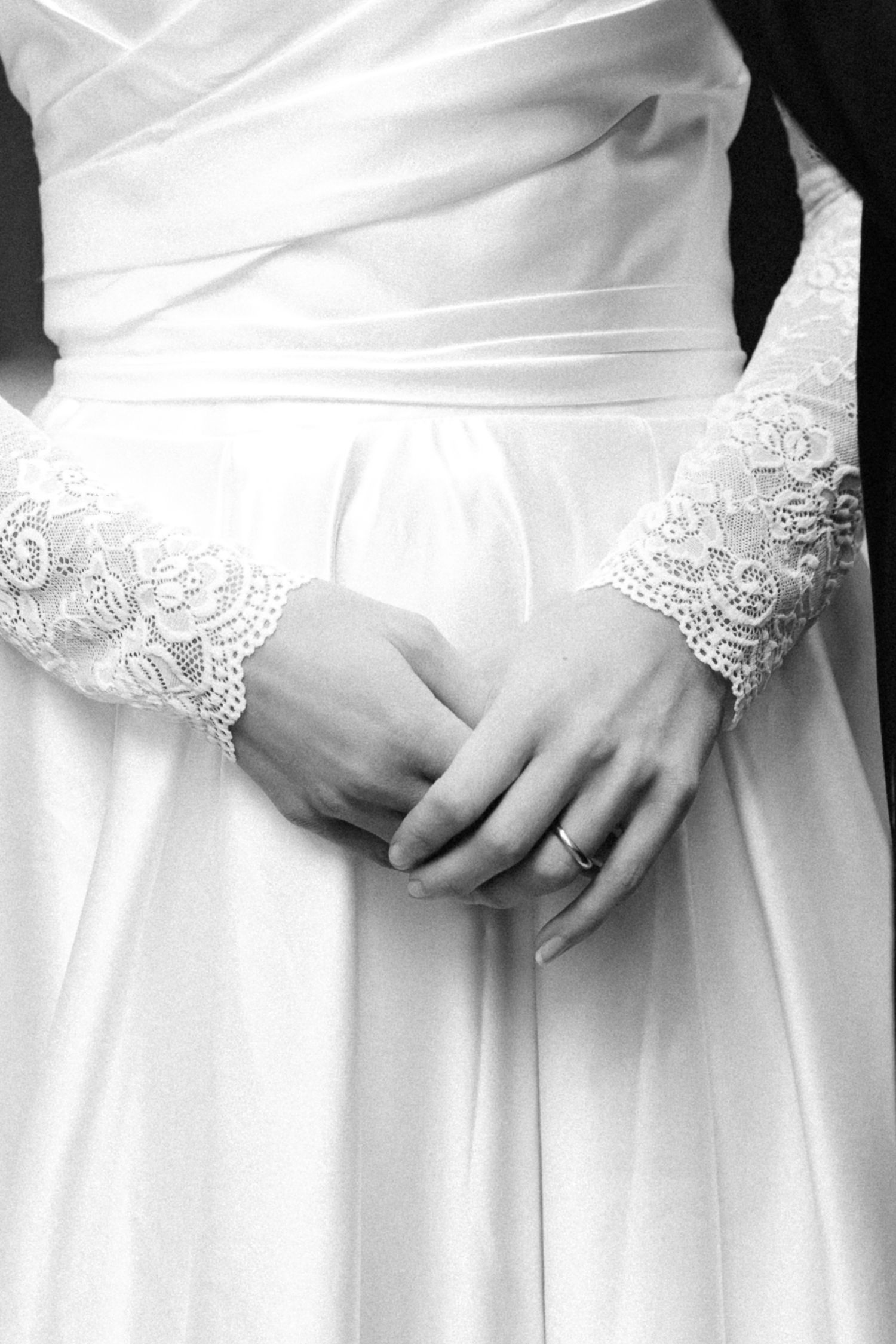 Black and white close-up of a bride's clasped hands with a ring, wearing a white satin wedding gown with delicate lace sleeves.