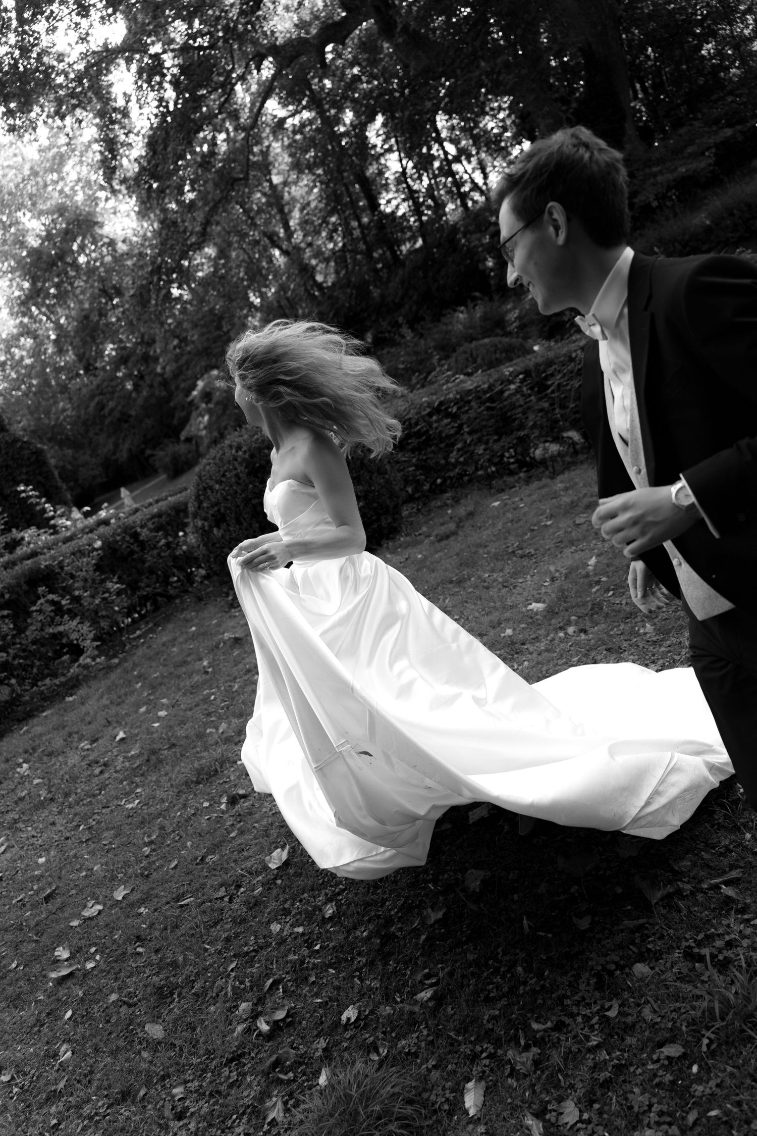 Black and white photo of a bride in a flowing gown twirling on a leafy lawn as a groom in a dark suit and glasses watches nearby.