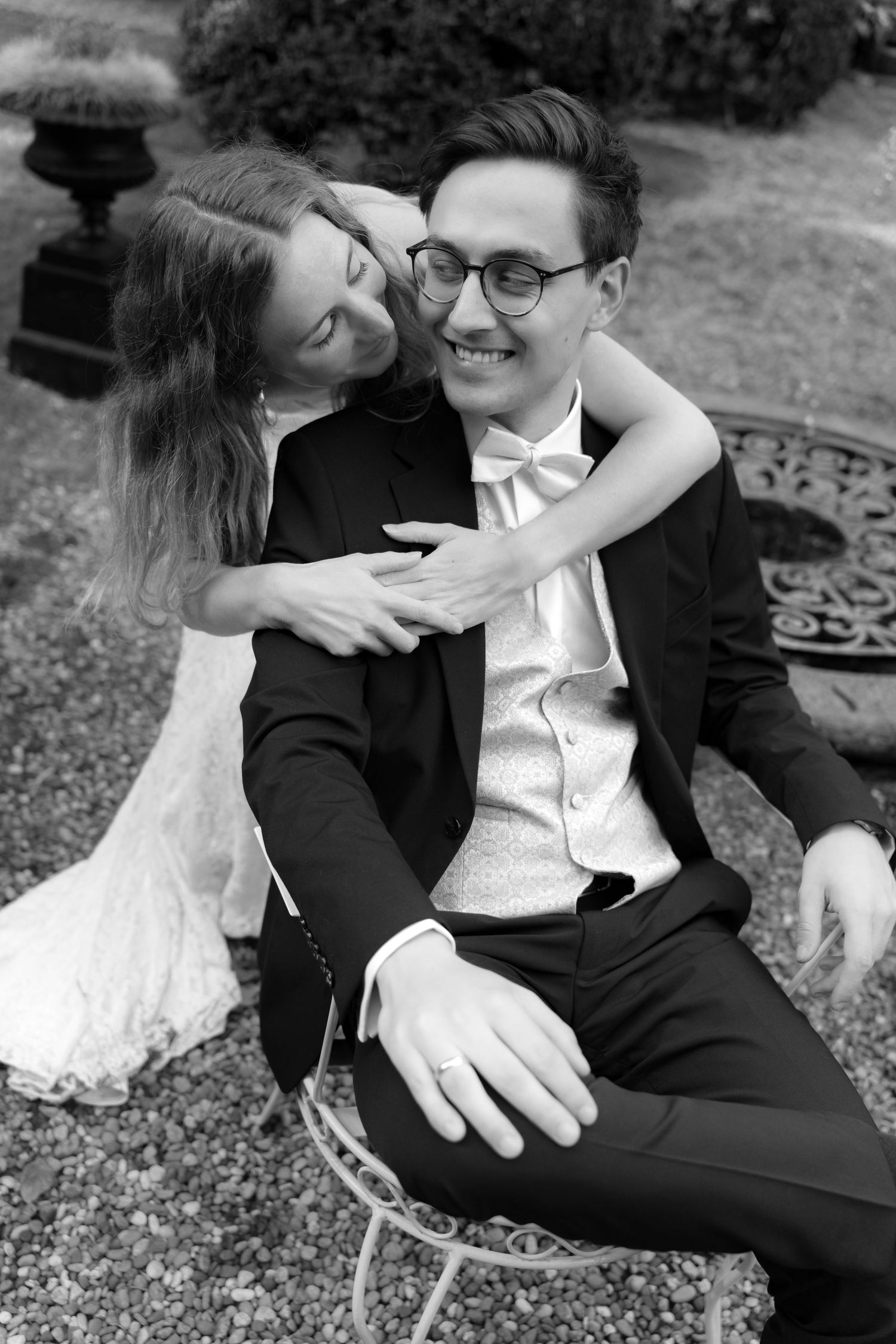 Black and white wedding portrait of a bride in a lace gown embracing a smiling groom in glasses and bow tie from behind in a garden.