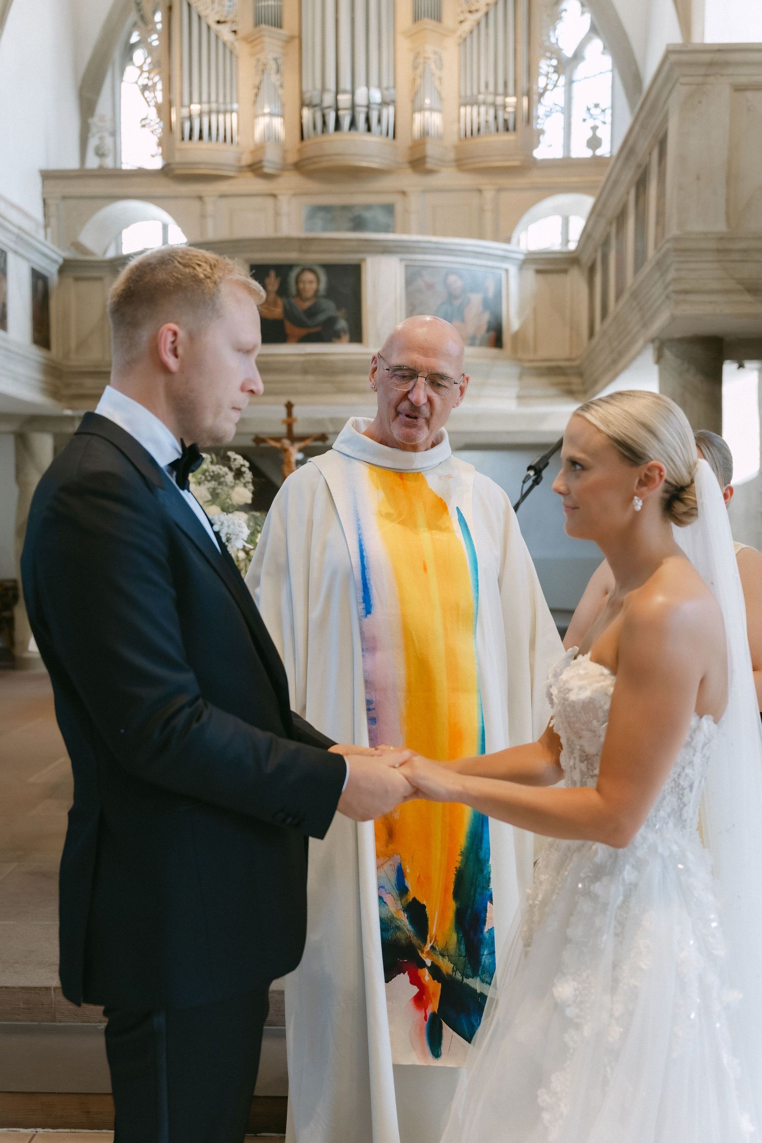 Bride and groom join hands during a church wedding ceremony as a priest in a white and colorful stole officiates before an ornate organ.