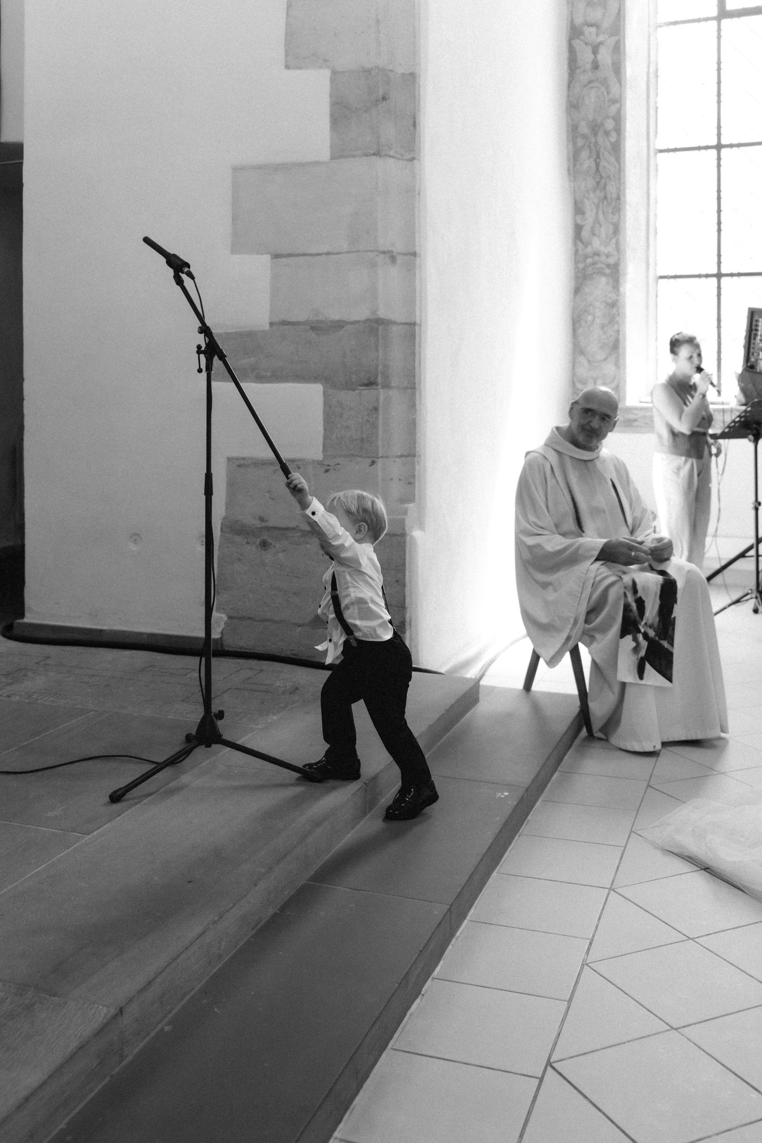 Black and white photo of a toddler reaching up to a tilted microphone stand in a stone church while a seated priest in robes smiles.