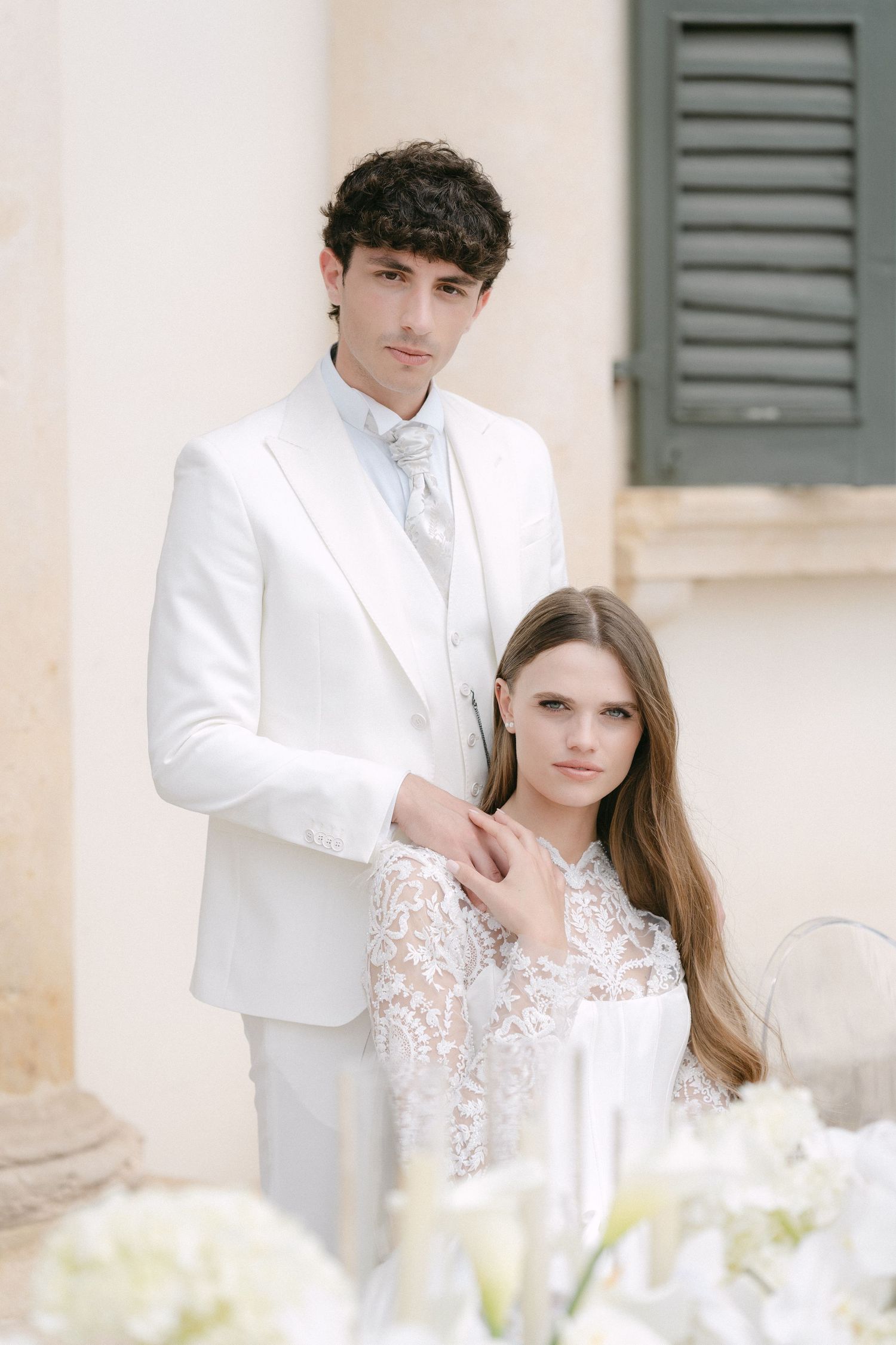 A couple in elegant white wedding attire poses together in front of a light colored building with green shutters. Villa Mosconi Bertani